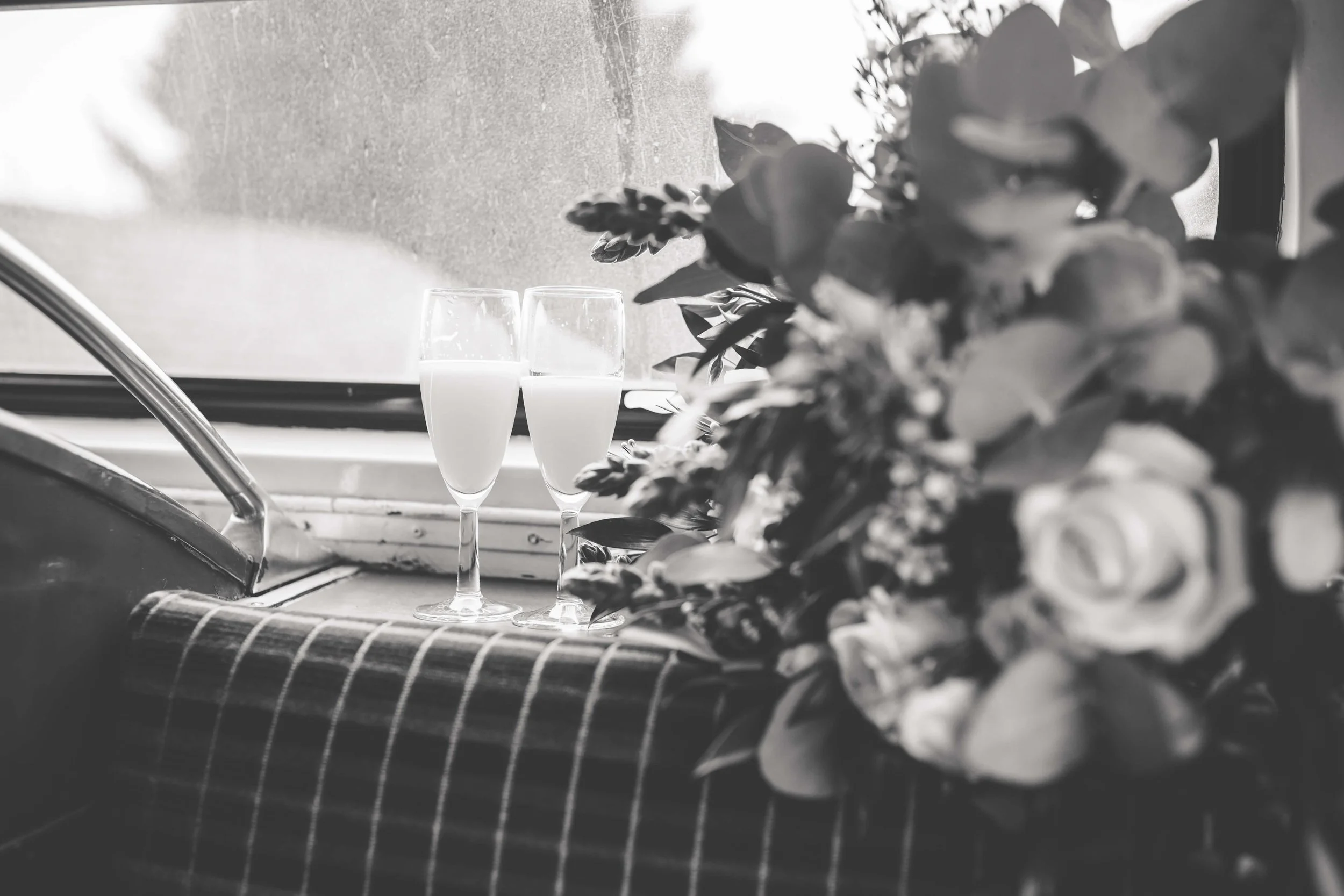 Two champagne glasses filled with a white beverage on a checkered cloth, next to a bouquet of flowers by a window.
