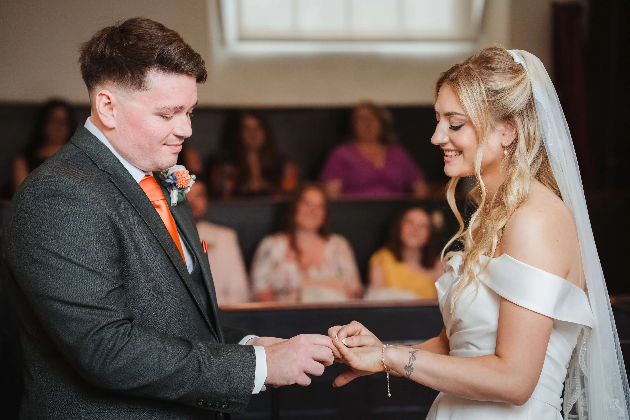 A bride and groom exchanging rings during their wedding ceremony indoors, with guests blurred in the background.