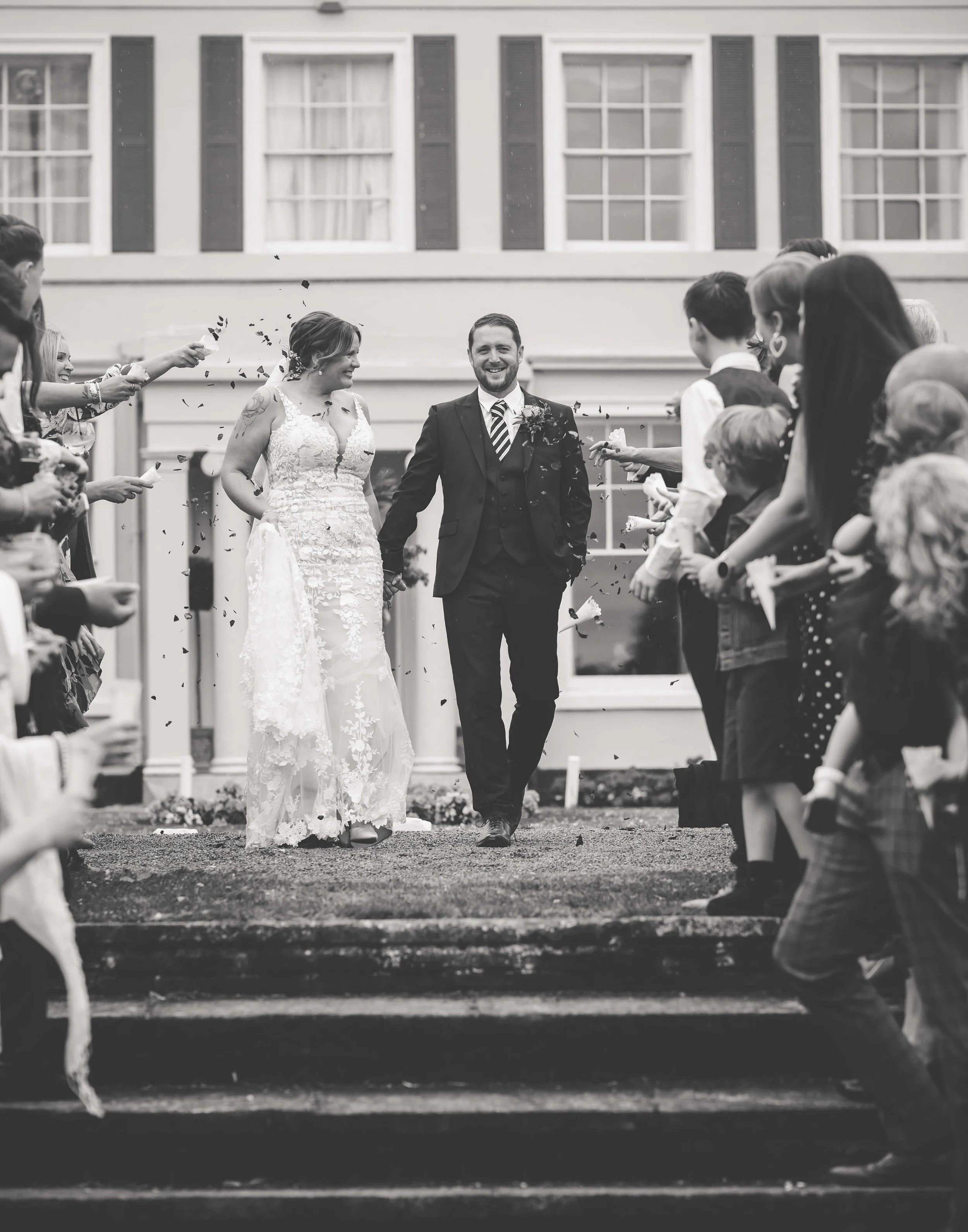 A wedding couple, a bride in a lace wedding gown and a groom in a suit, walking down steps surrounded by celebrating friends and family. The scene is joyful, with confetti in the air, outside a building with large windows.