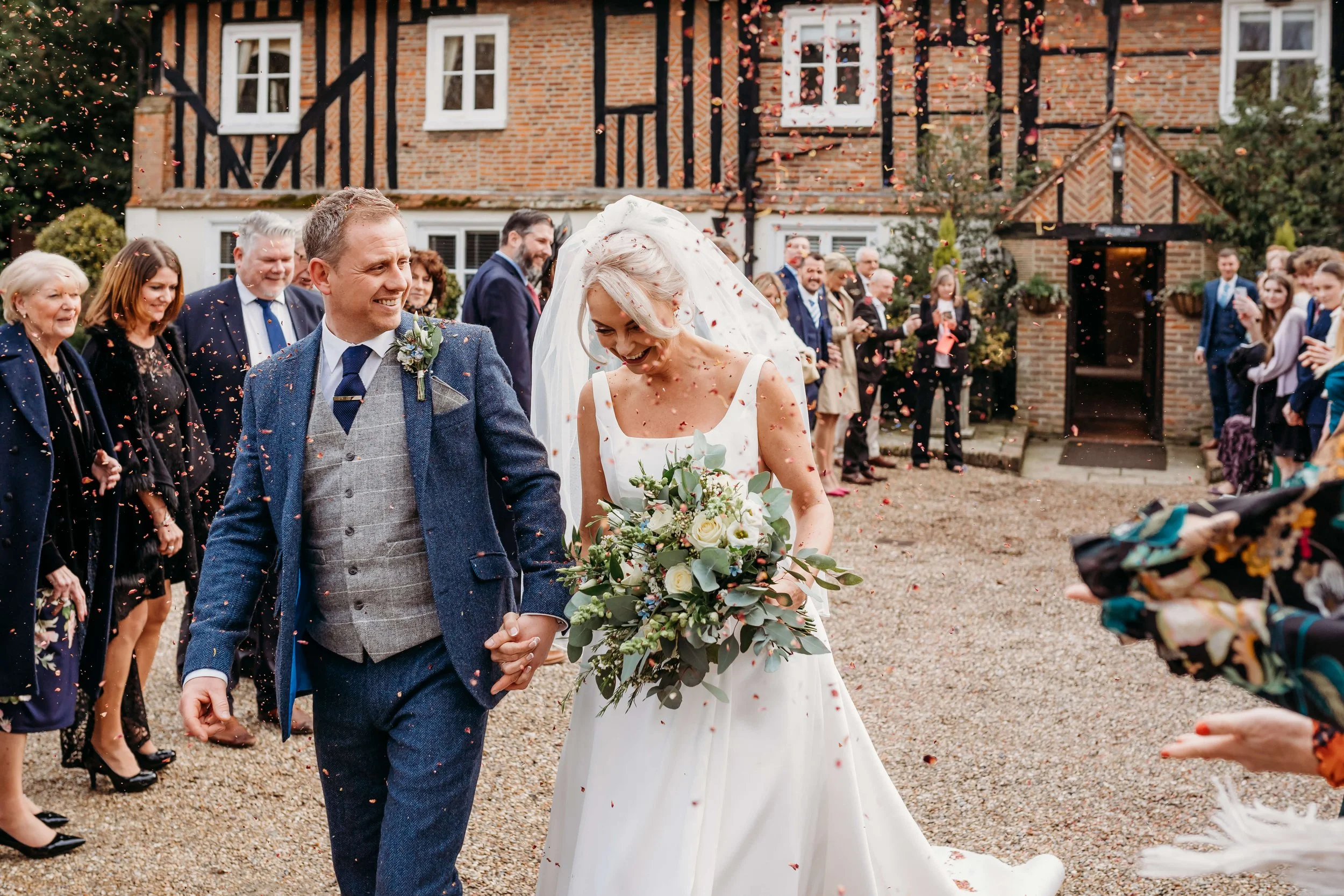 A bride and groom holding hands and smiling at each other as wedding guests throw confetti behind them outside a brick and timber-framed building.