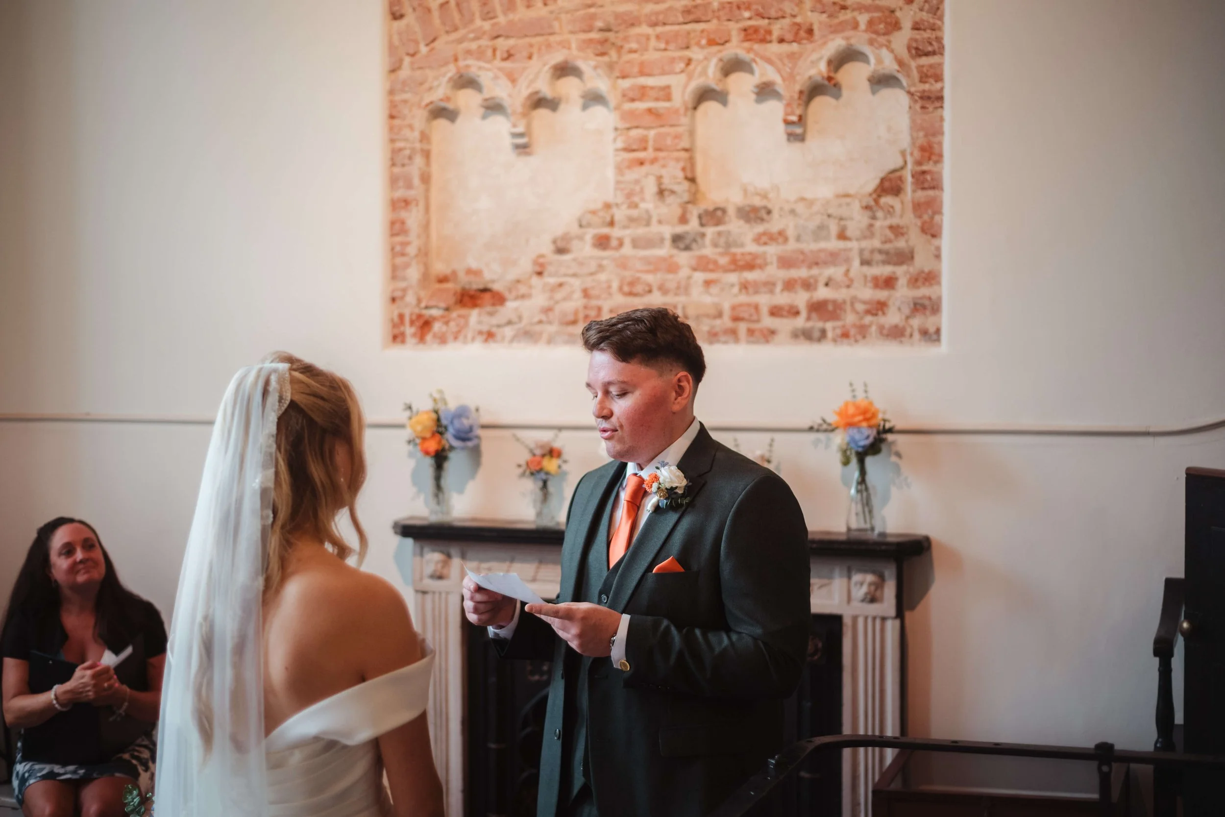 A bride and groom exchange vows during a wedding ceremony indoors with guests watching. The groom is reading from a paper, dressed in a dark suit with an orange tie and boutonniere, the bride is wearing a white off-the-shoulder dress and a veil. In t