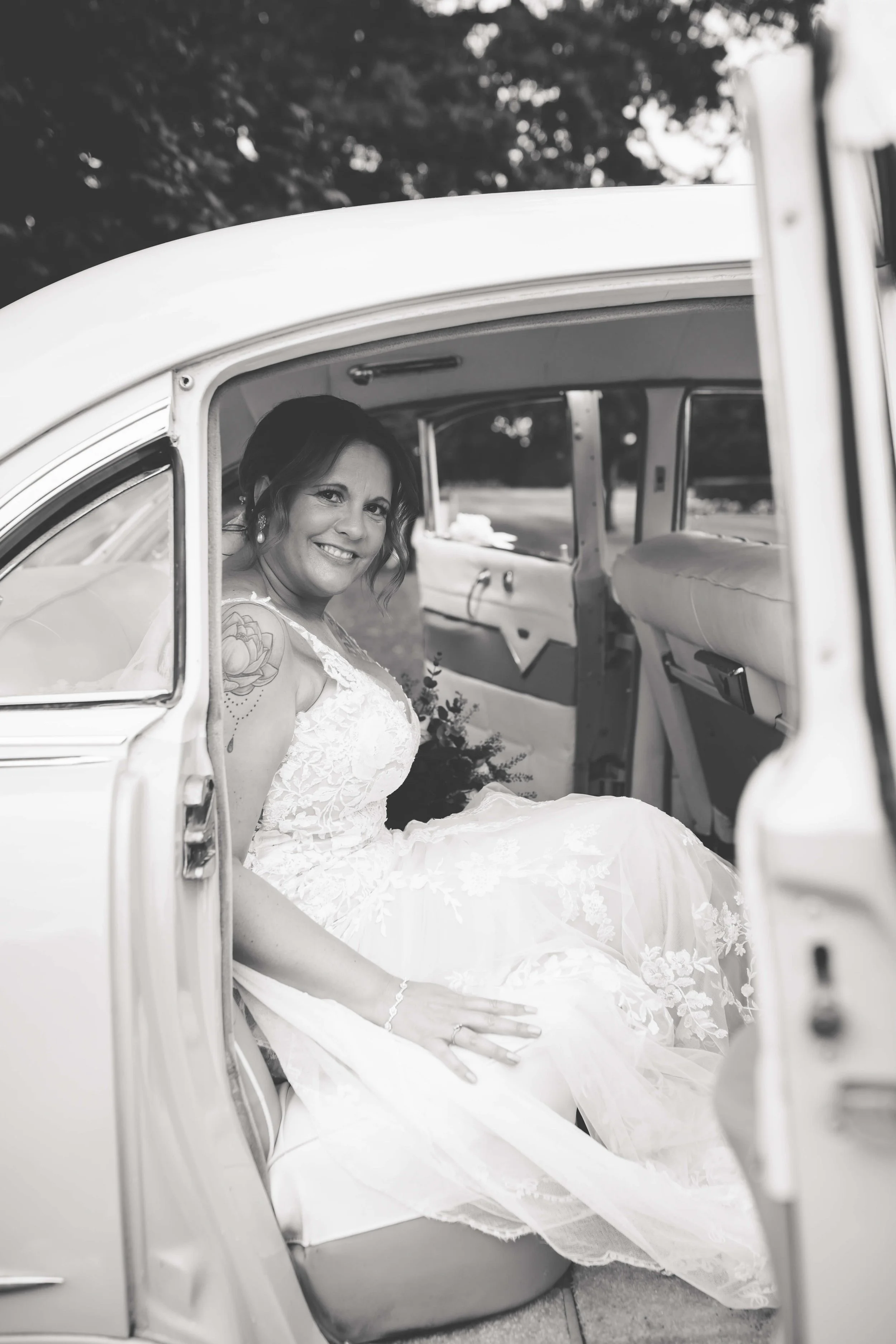 A woman in a wedding dress sitting inside a vintage car, looking out and smiling.