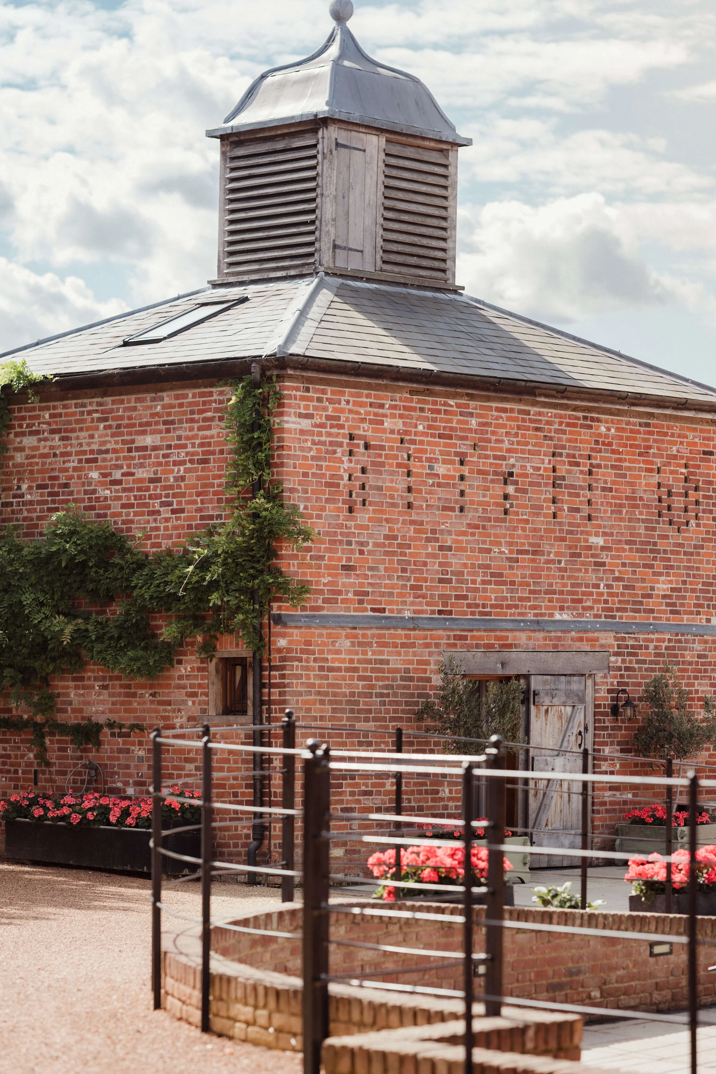 A red brick barn with a gray metal roof and a small wooden cupola on top. There are flowering plants and a fenced patio area in front of the barn.