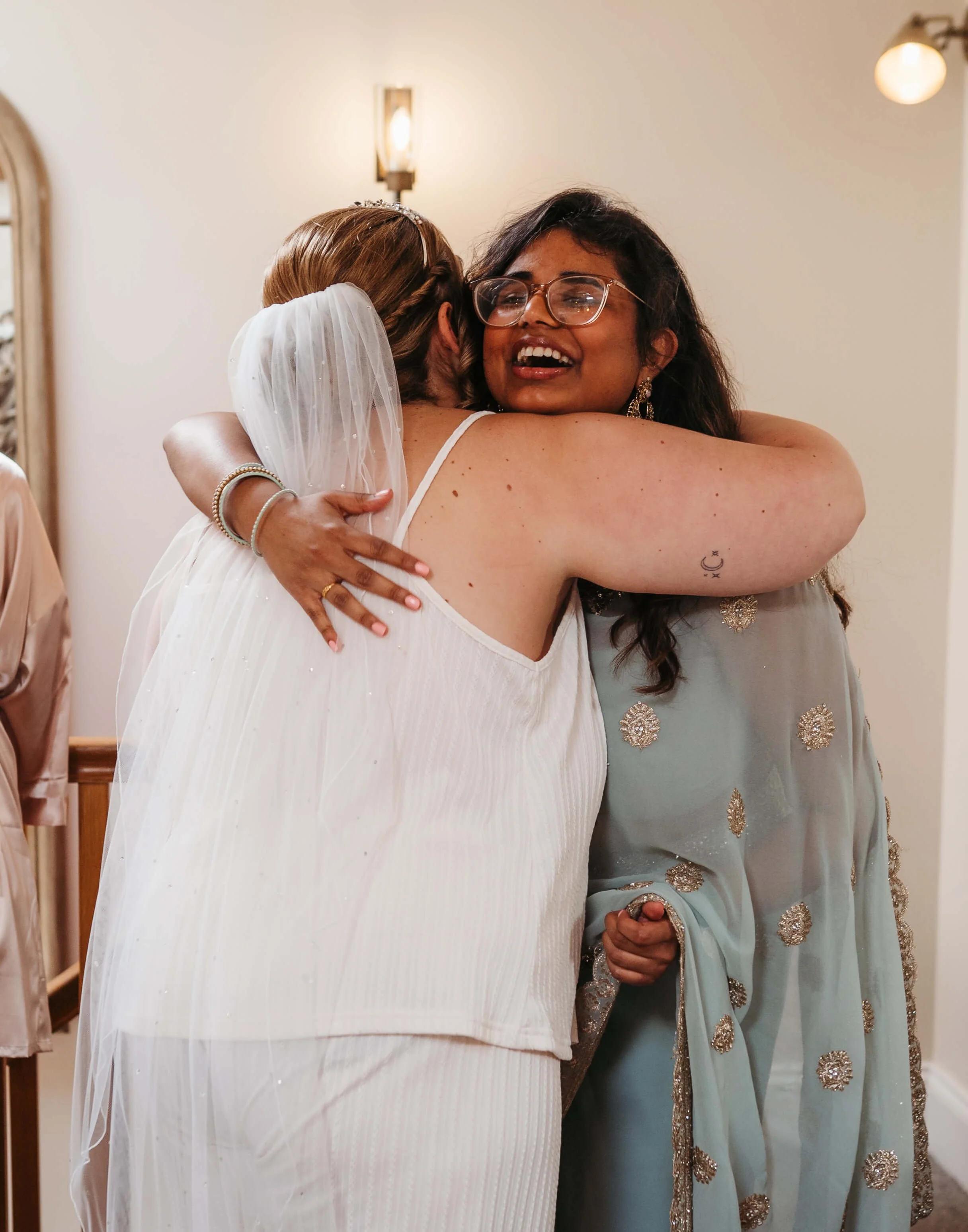 Two women hugging, one in a white wedding dress and veil, and the other in a light blue traditional outfit with gold embellishments, sharing a joyful moment.