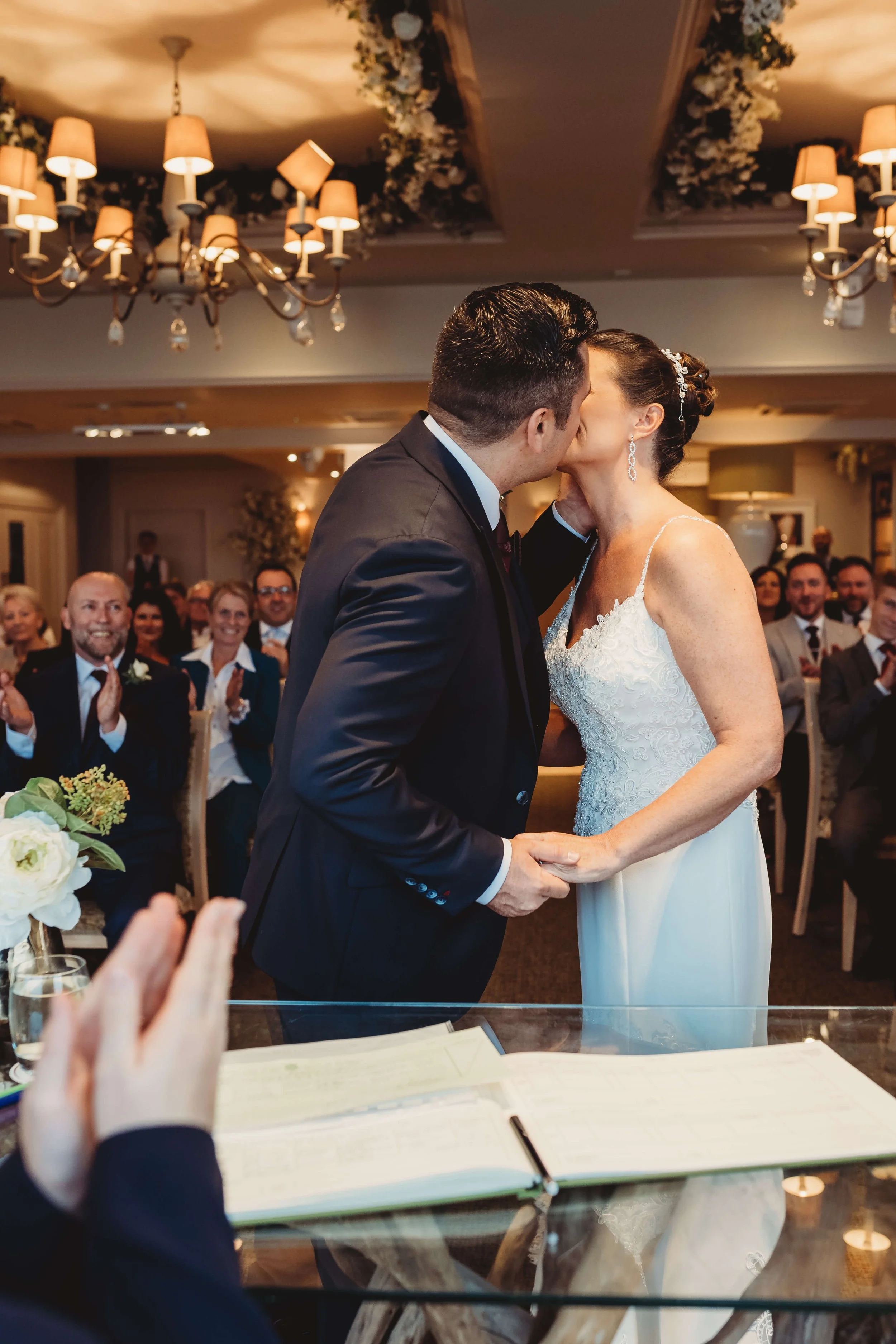 A newlywed couple sharing a kiss during their wedding ceremony, surrounded by seated guests in a decorated reception hall.