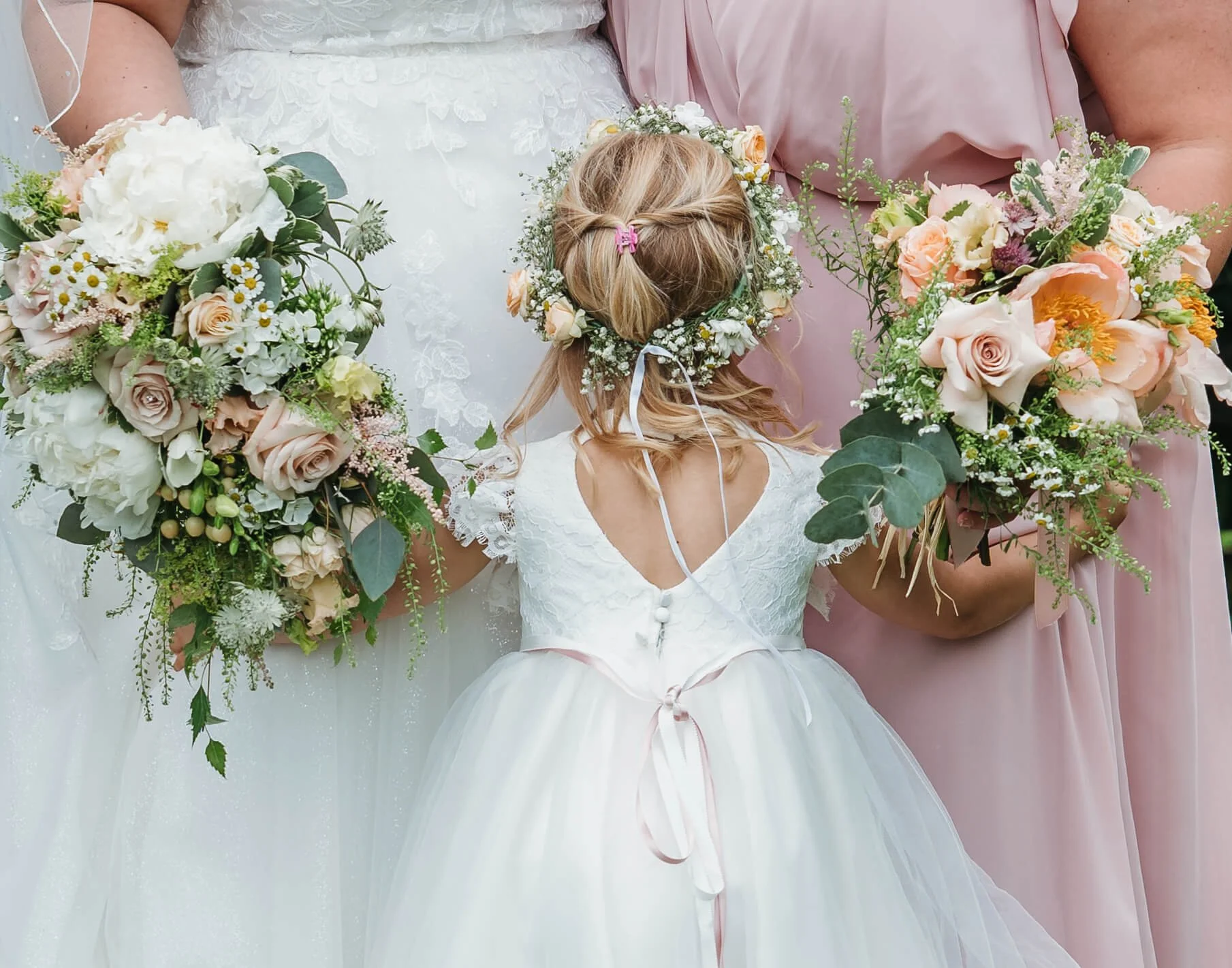 A young girl in a white dress holding the hands of two women in pink dresses, all holding bouquets of flowers at a wedding.