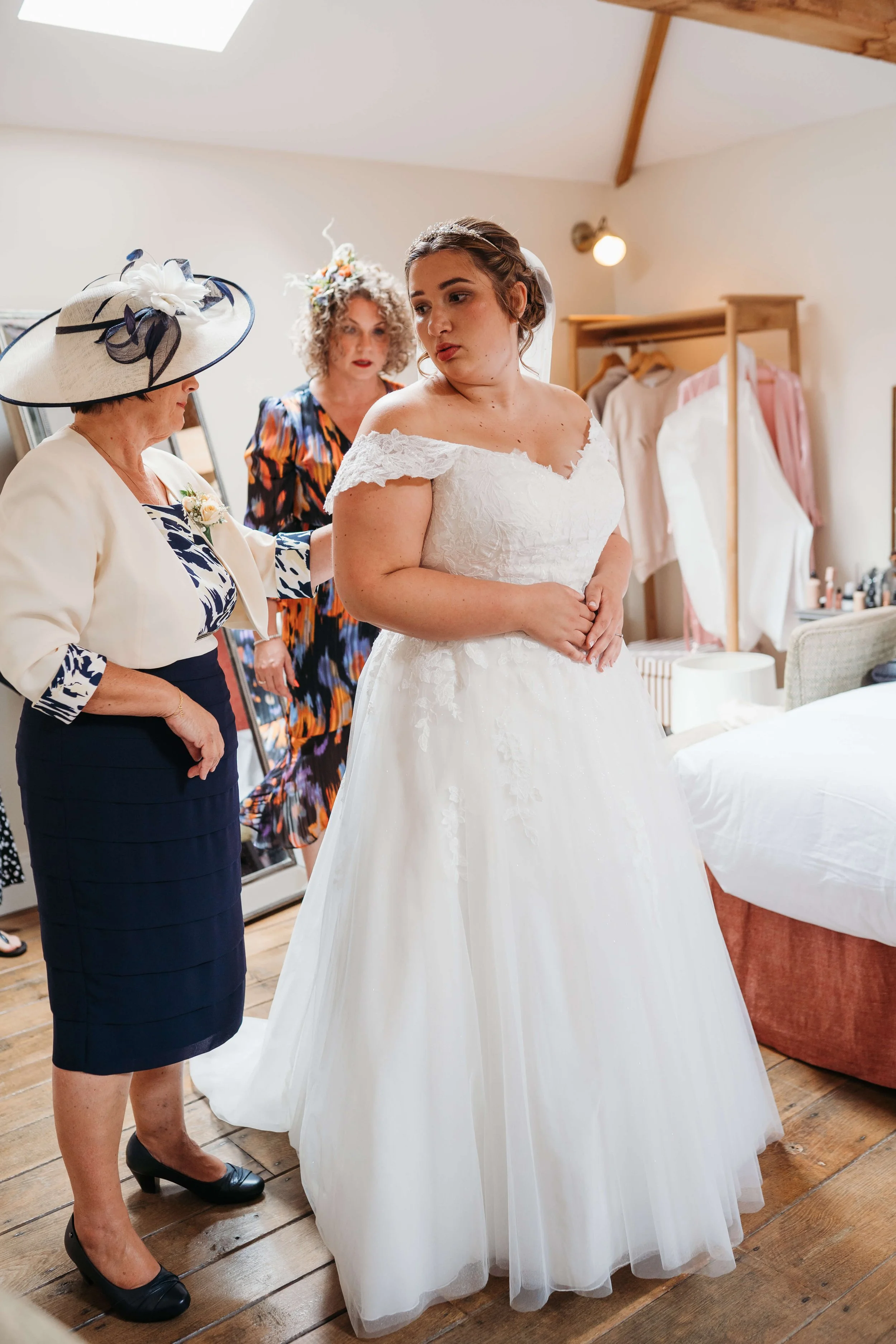 A bride in a white wedding gown, standing in a bedroom with her mother and an older woman, possibly a family member or friend, helping her prepare for her wedding. The room has wooden floors, a clothing rack with dresses, and a bed.