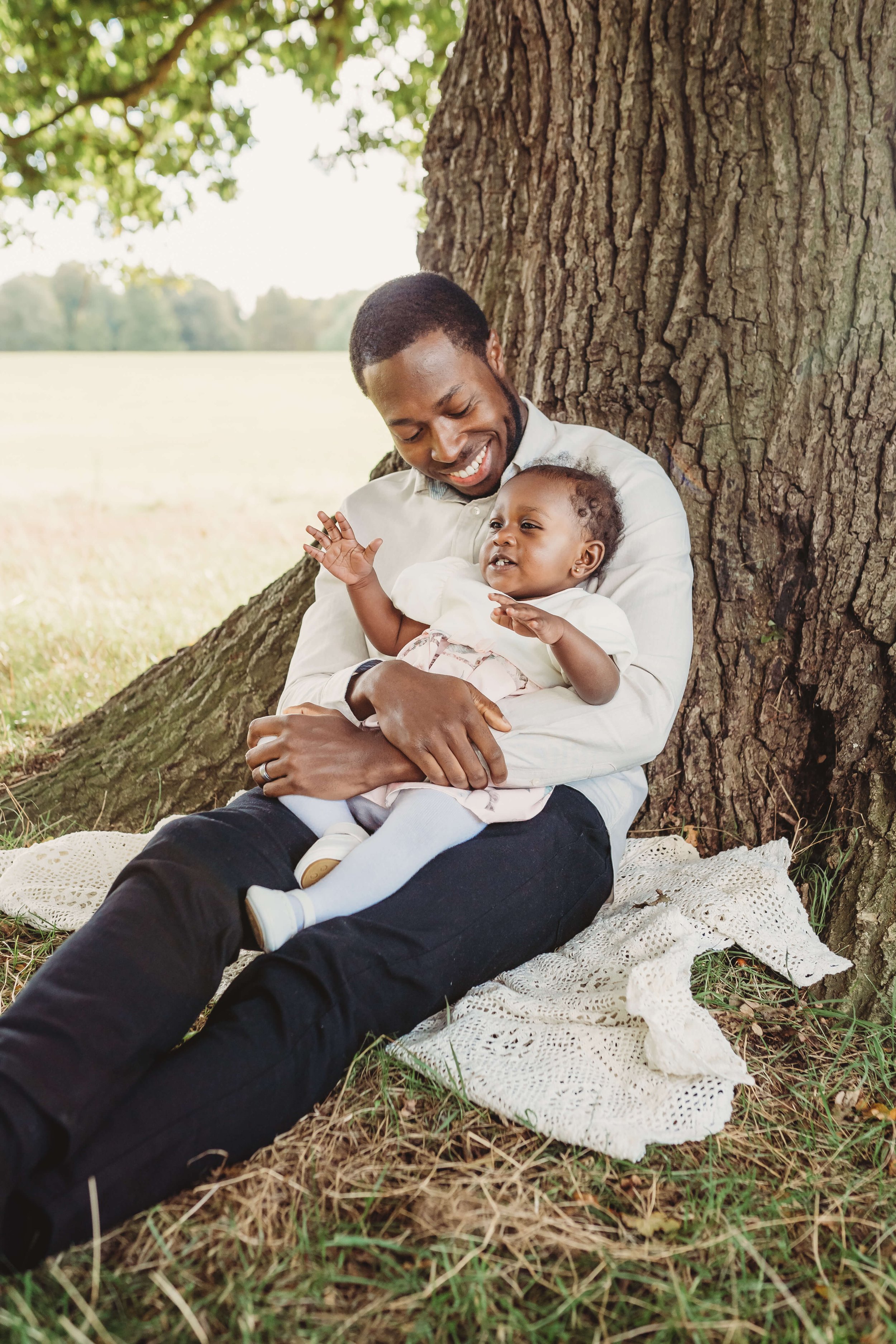 A man and a young girl sitting on a blanket under a large tree outdoors, smiling and engaging with each other.