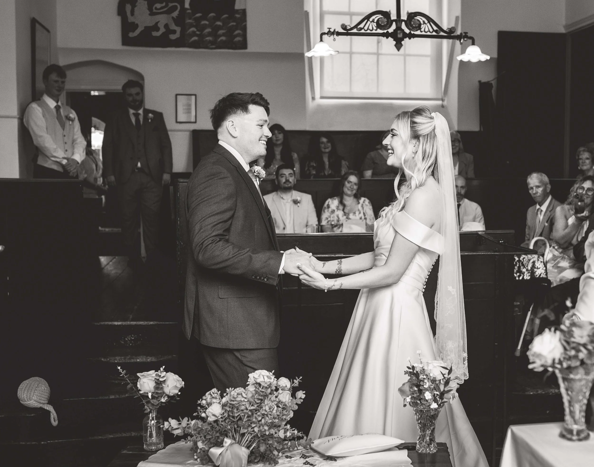 A bride and groom holding hands during their wedding ceremony, smiling at each other, with guests in the background.