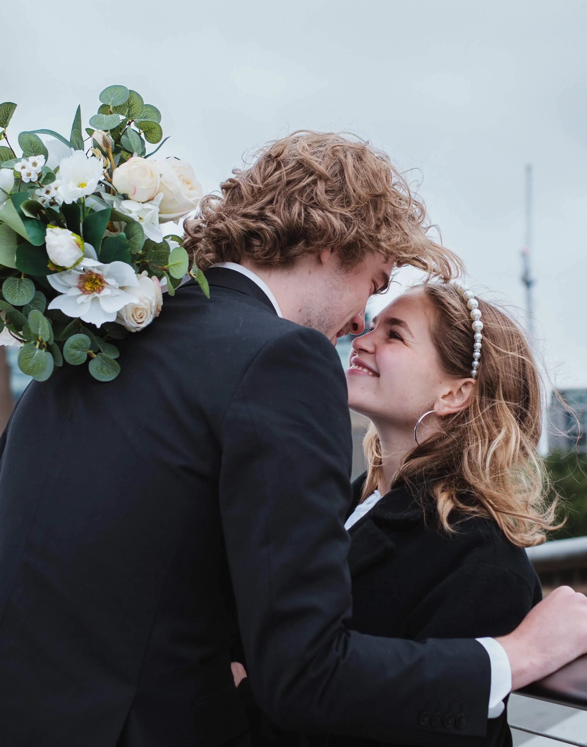 A couple is embracing closely outdoors, with the man carrying a bouquet of white roses and green foliage on his shoulder. They are gazing into each other's eyes, smiling, with the sky and some structures in the background.