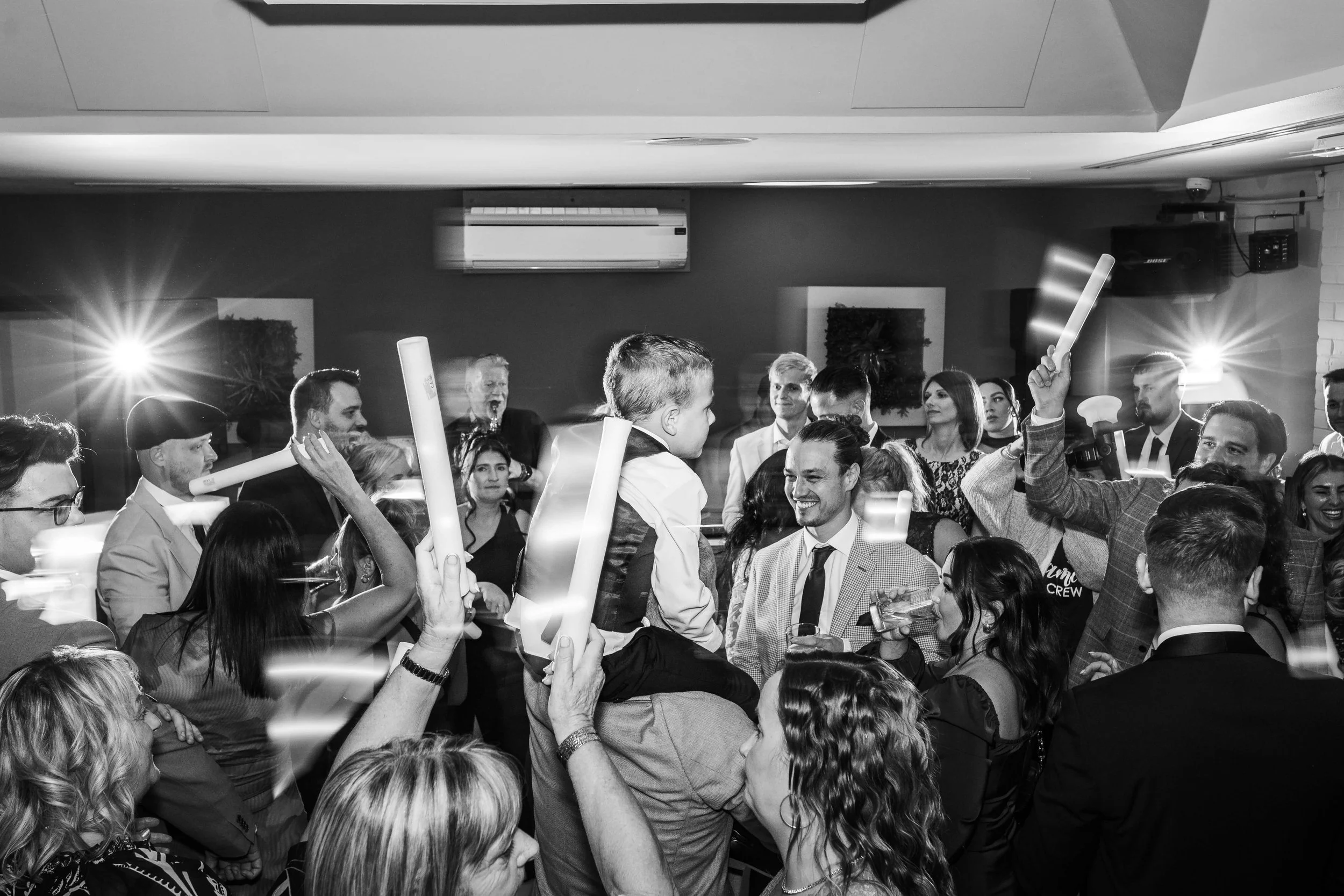 Guests celebrating at a lively party or event, with a man on someone's shoulders, surrounded by smiling people holding glow sticks and drinks.