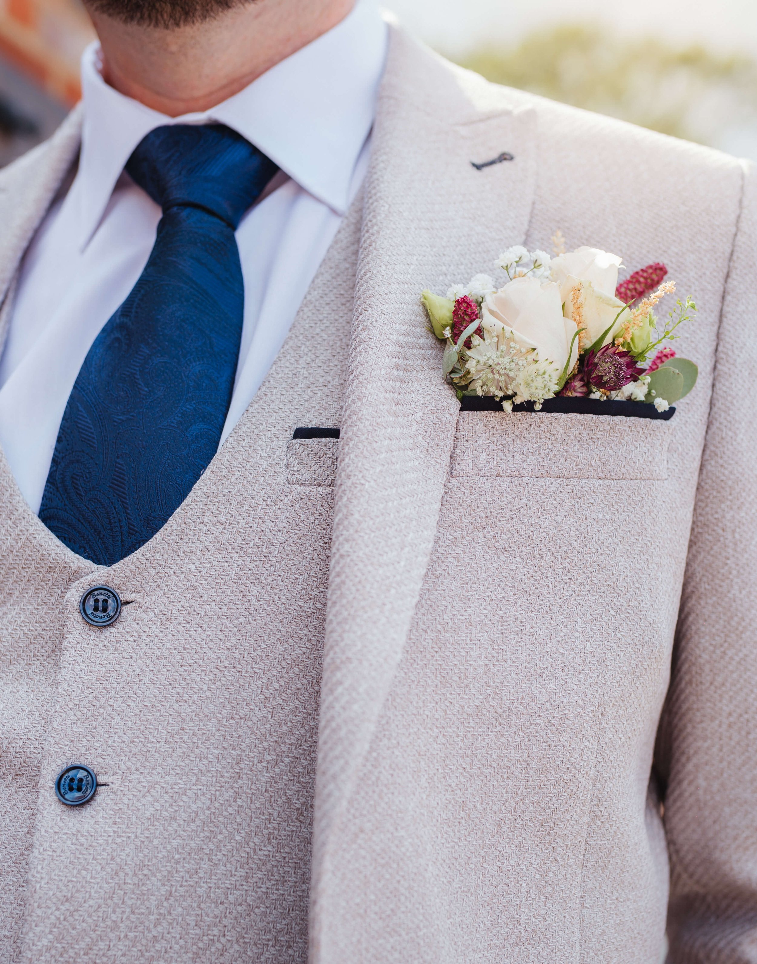 Close-up of a man wearing a beige suit with a boutonniere of white and pink flowers on the left lapel, a white shirt, and a navy blue tie with a paisley pattern.