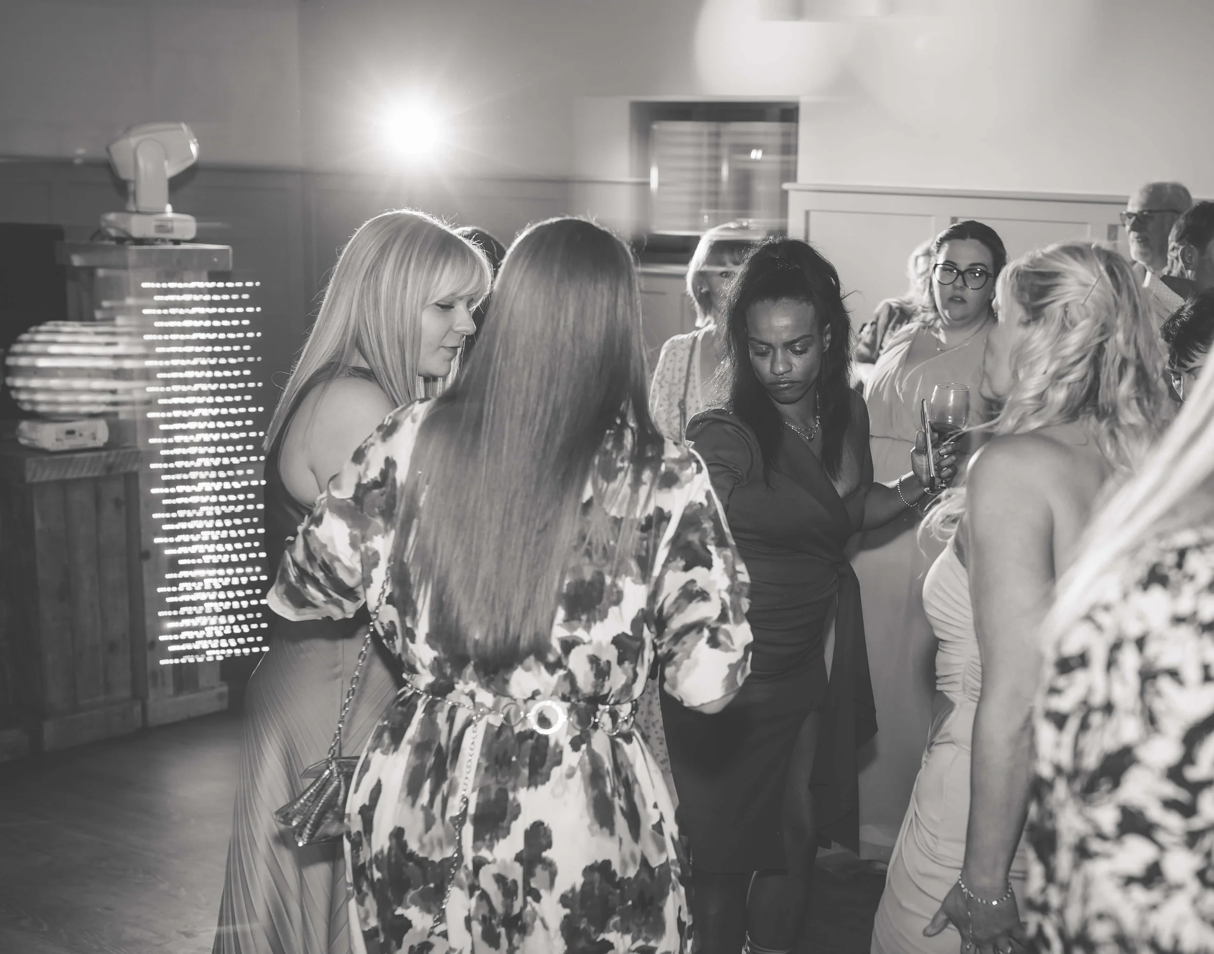 Group of women gathered at a social event, some holding glasses, engaged in conversation, indoors with a bright light source in the background.