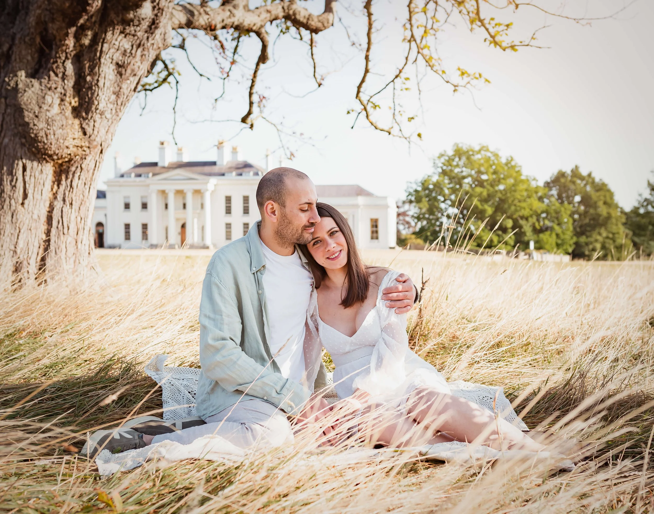 A young couple sitting on a blanket in a field of tall grass with a white mansion in the background, sharing a tender moment outdoors.