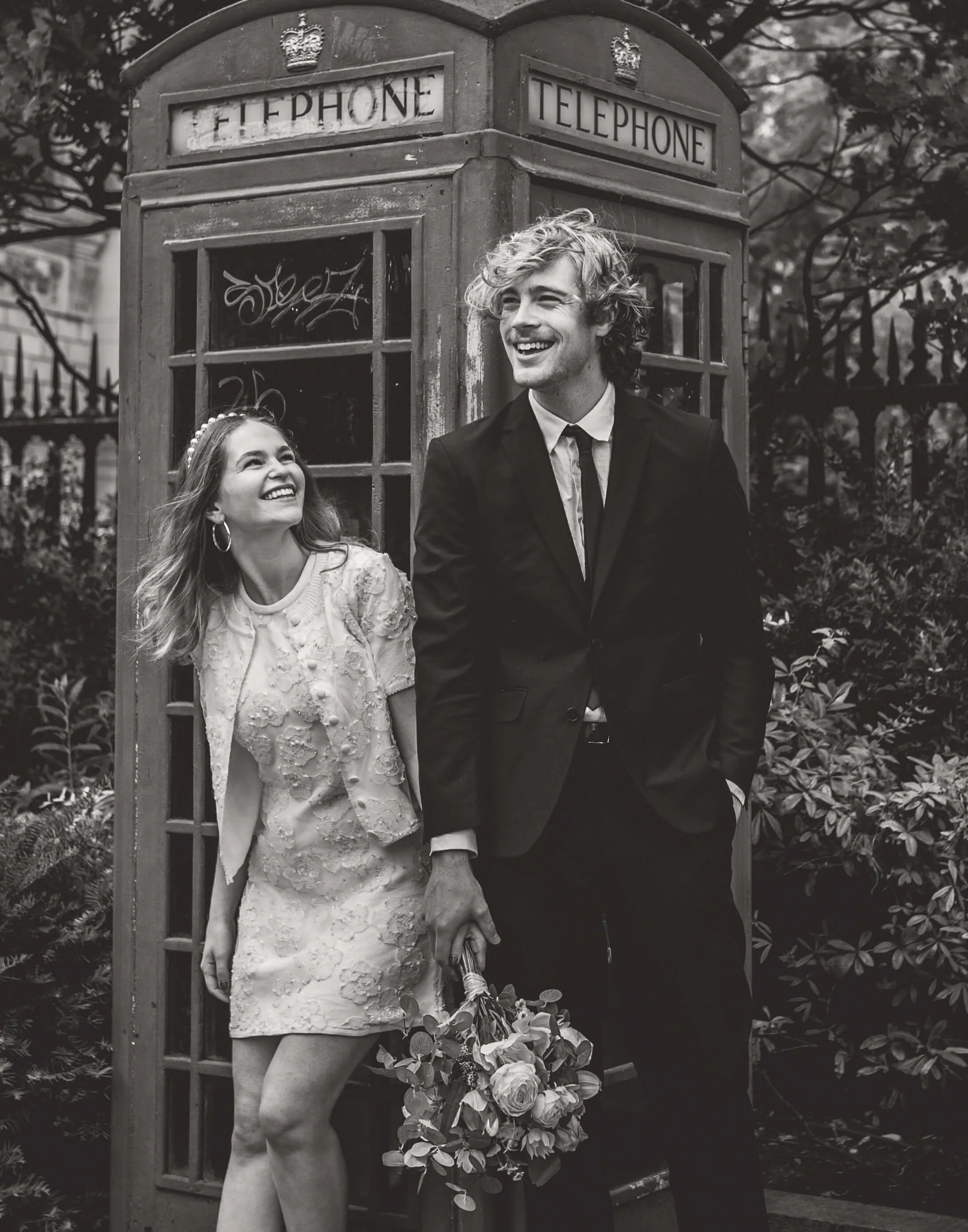 A smiling couple in wedding attire standing in front of a vintage telephone booth, with the woman holding a bouquet and the man holding her hand.