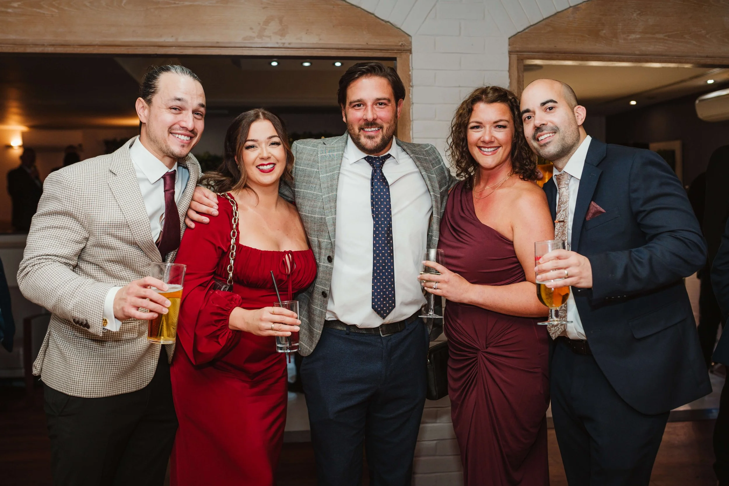 Group of six adults at a social gathering, smiling, holding drinks, standing close together in an indoor setting.