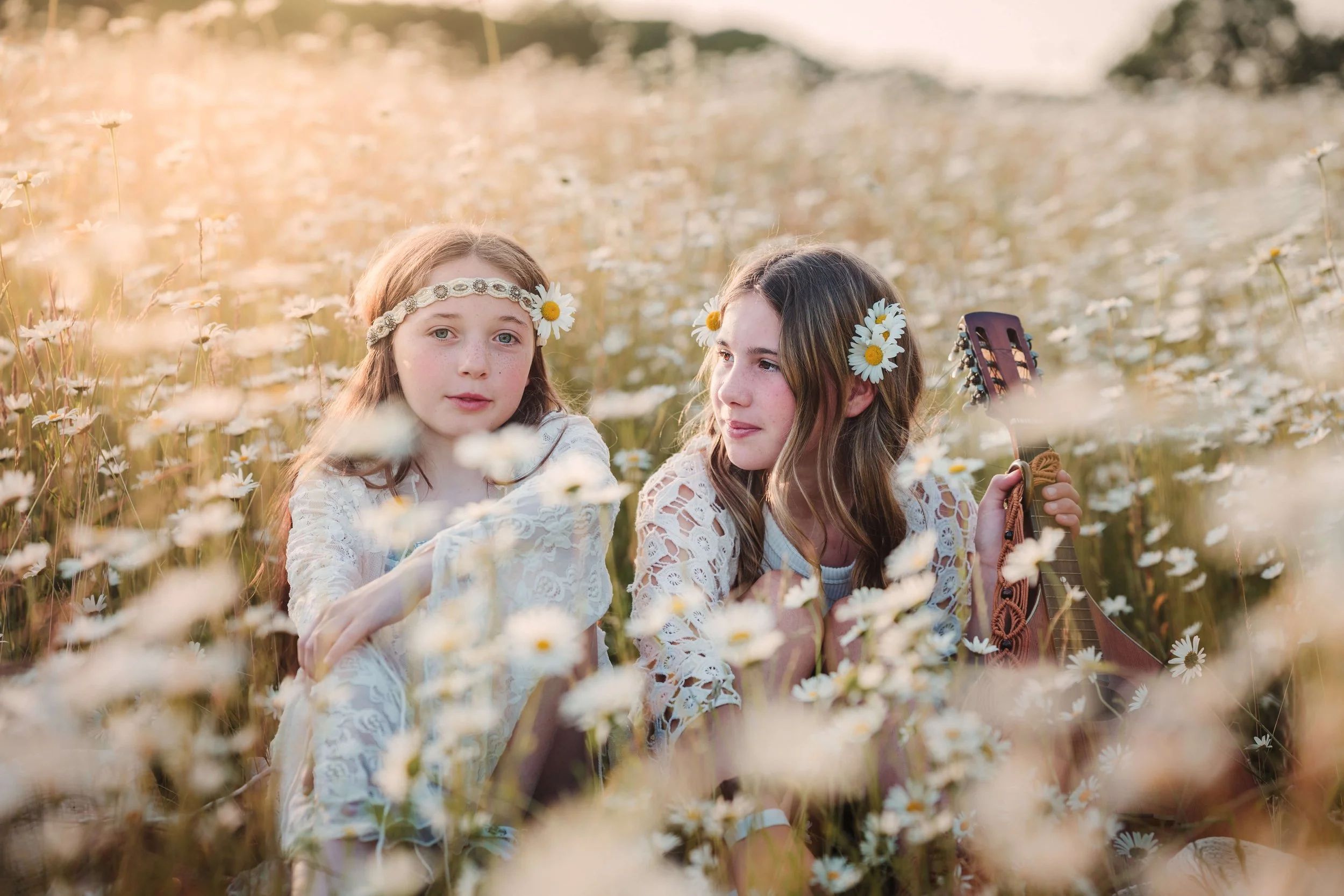 Two young girls with daisy flowers in their hair sitting in a field of daisies during sunset, with one holding a guitar.