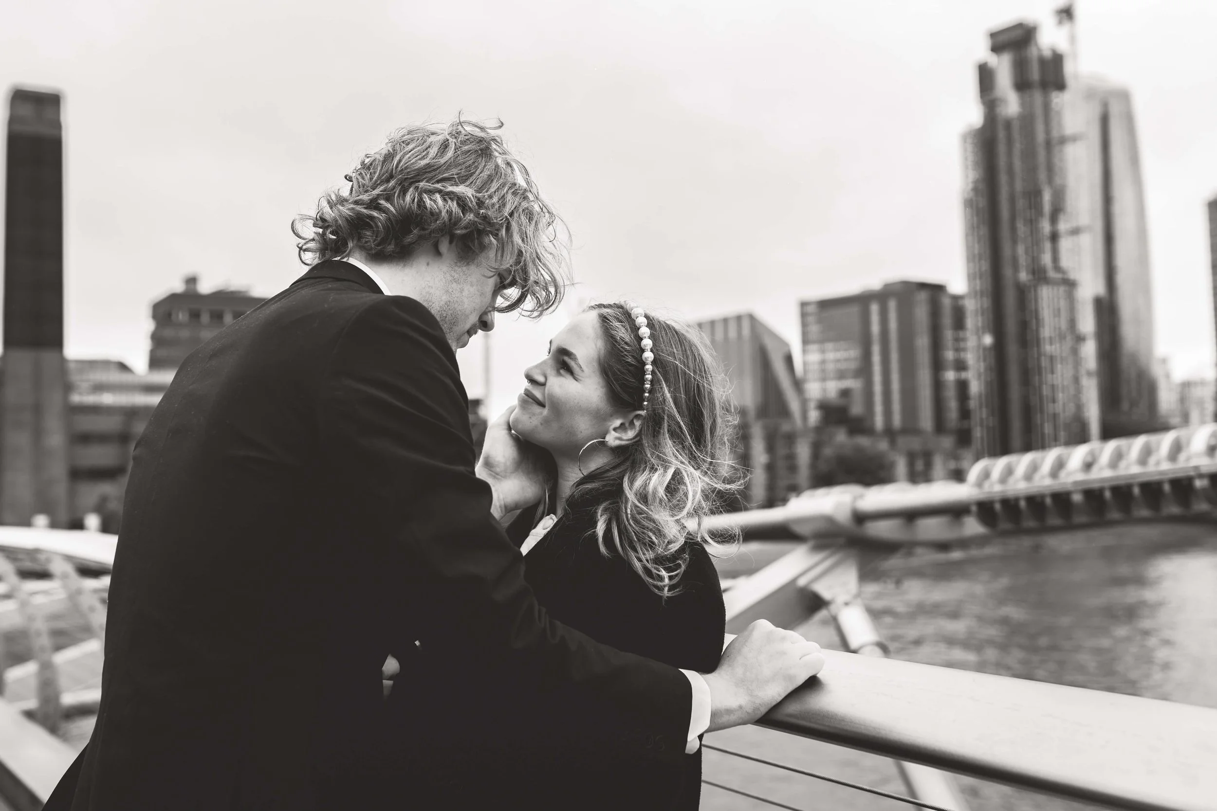 A black and white photograph of a couple on a bridge or walkway in a city, with tall buildings in the background. The man is leaning in toward the woman who is smiling and looking at him, with her hand resting on his chin.