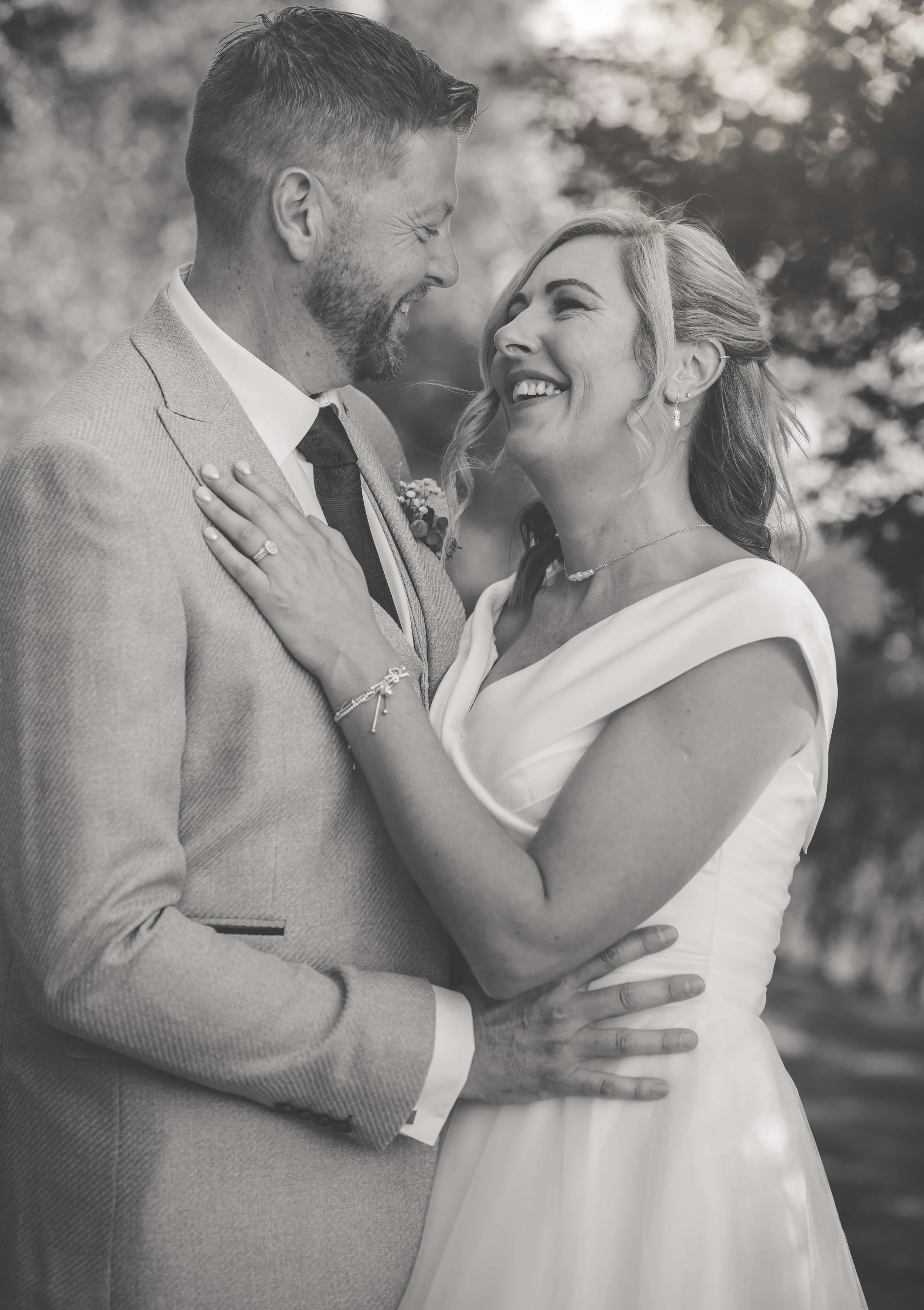 A black and white photo of a happy couple on their wedding day, gazing at each other and smiling, with a blurred outdoor background.
