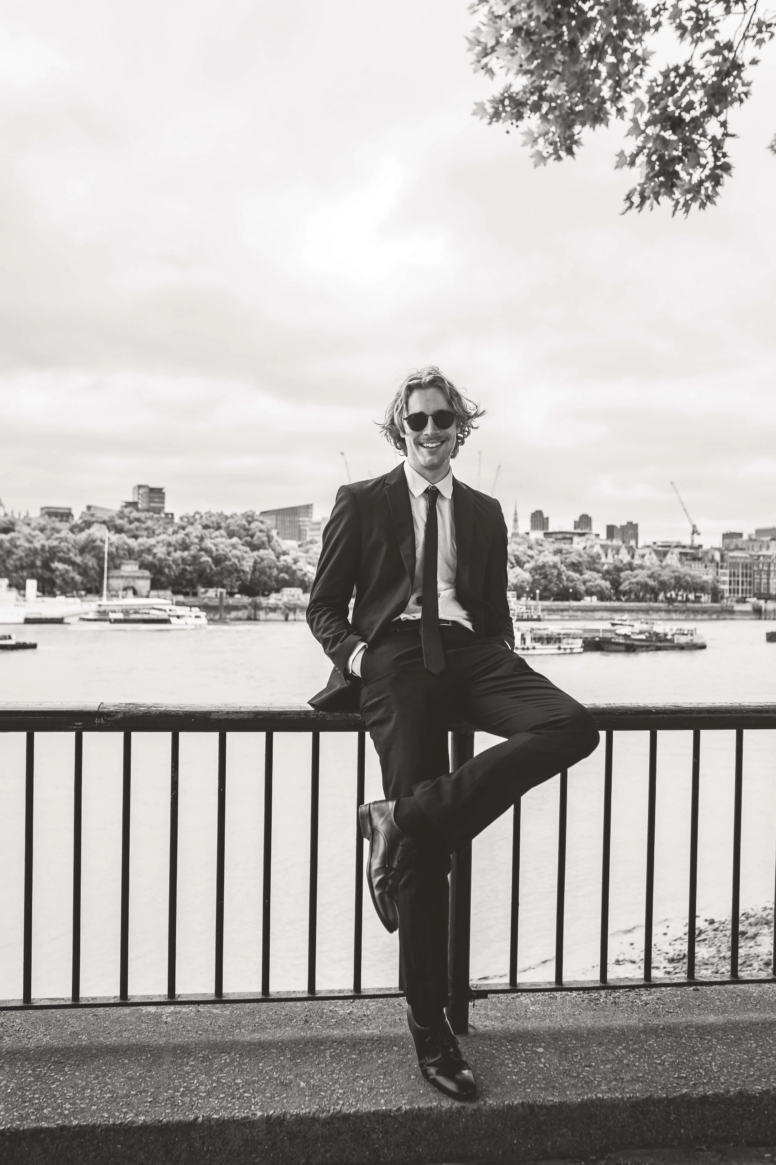 A man in a suit and sunglasses sitting on a railing near a body of water with a city skyline and trees in the background.