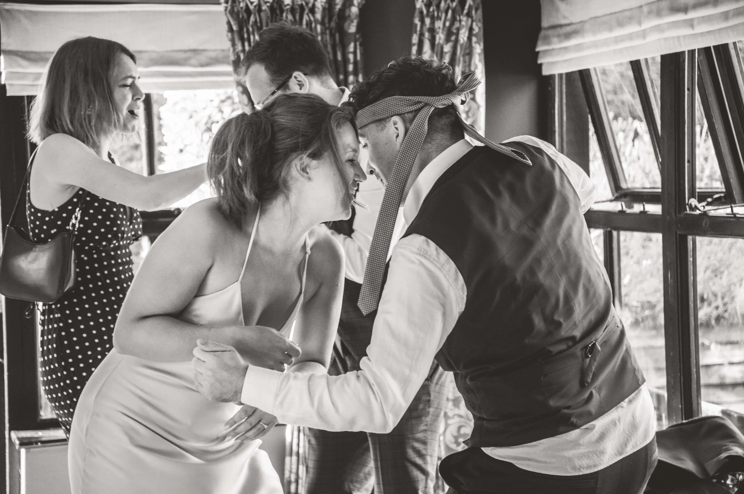 A woman in a wedding dress and a man with a bandana on his head are bowing their foreheads to each other, surrounded by friends. The scene is in a bright room next to windows.