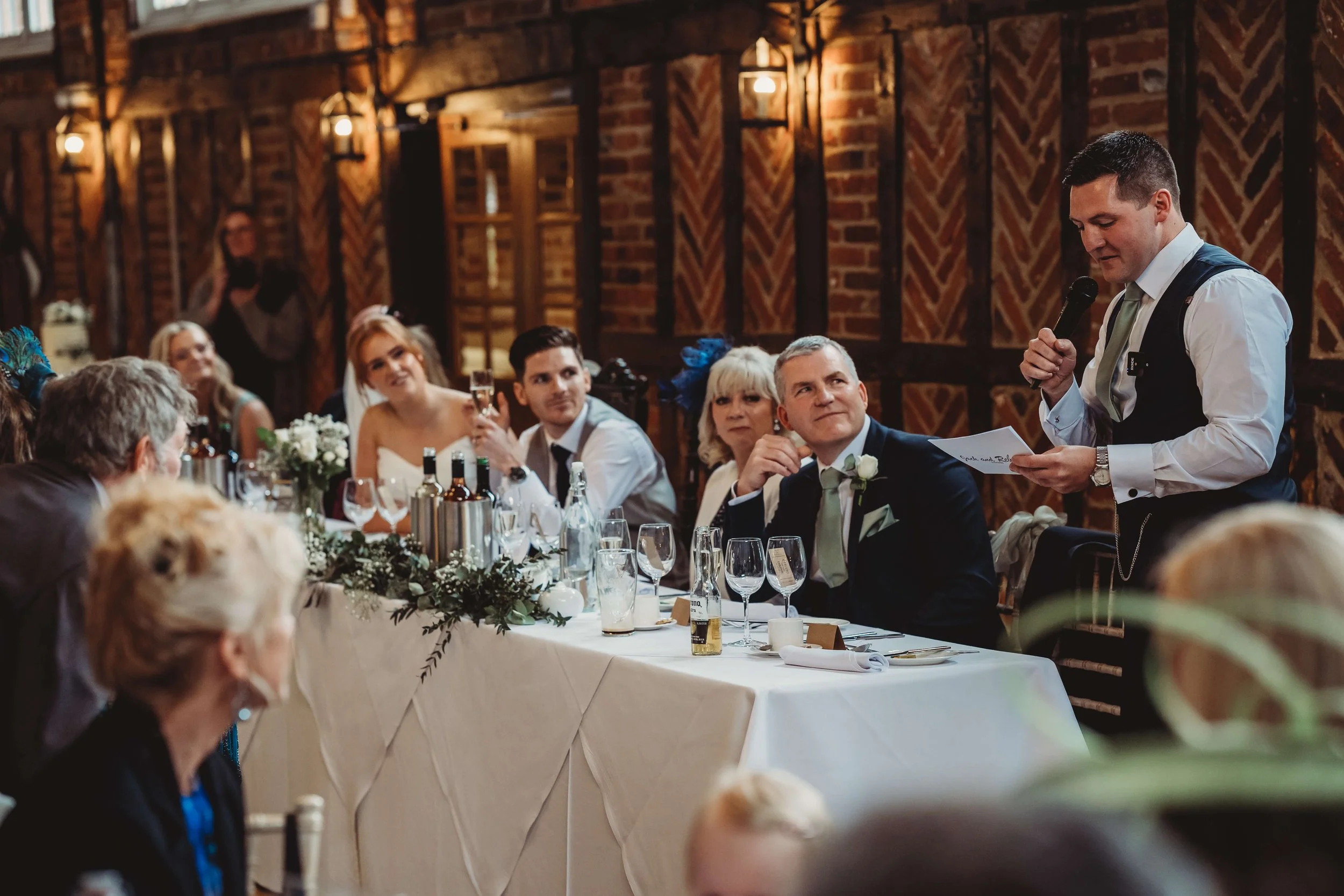 A man giving a speech at a wedding reception with guests seated at a long table listening attentively.