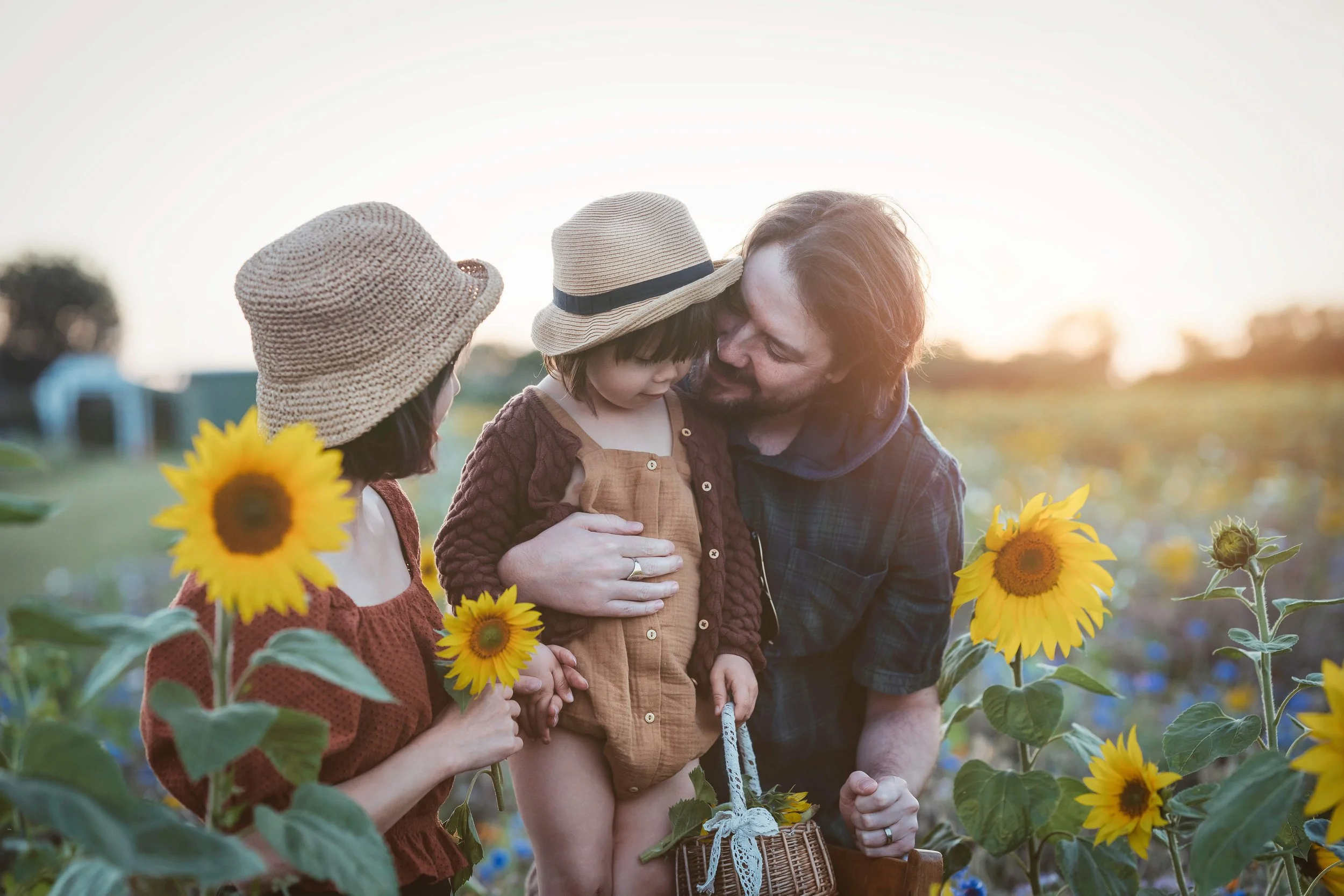 A family of three with two children, all wearing sun hats, standing among sunflowers during sunset. The father is holding the little girl, and the woman is holding a sunflower and engaging with them.