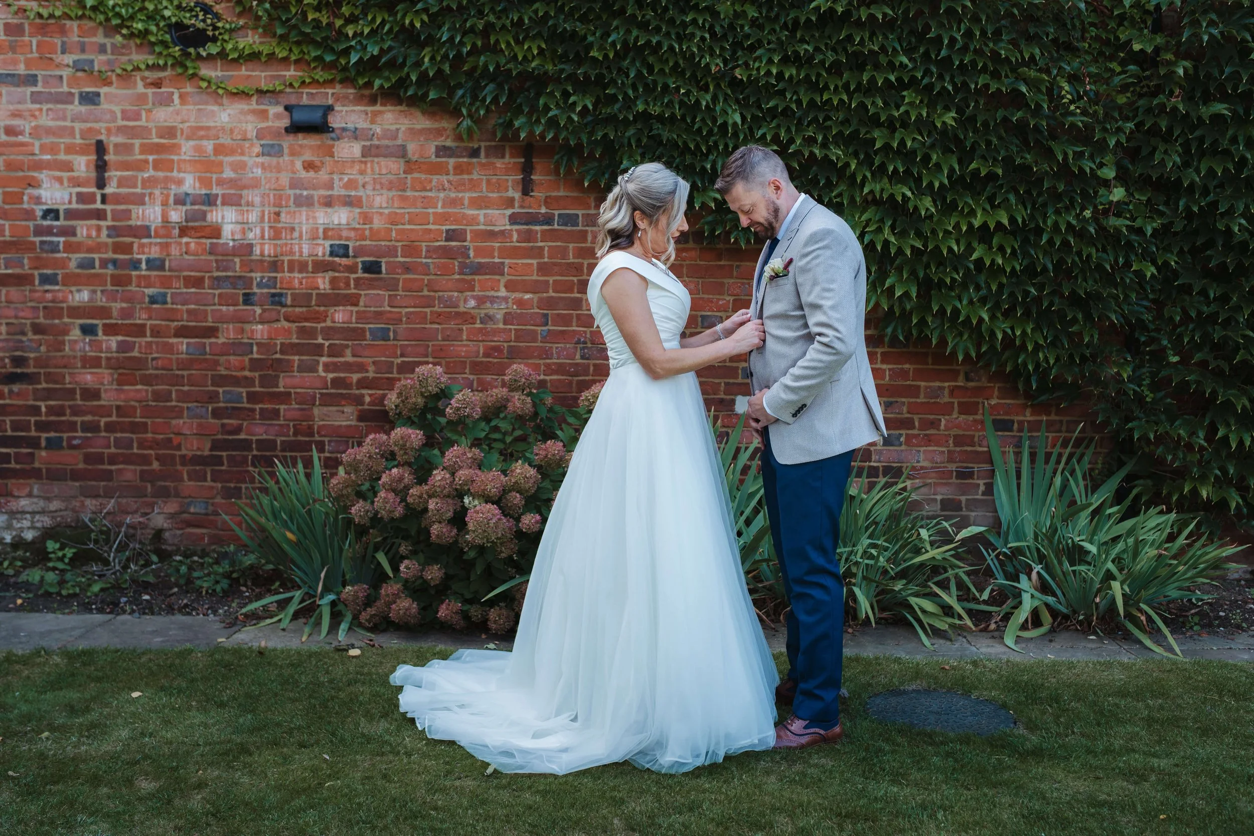 A bride in a white wedding dress and a groom in a gray blazer and blue pants standing close together outside against a brick wall covered in green ivy. The bride is pinning a boutonnière on the groom's blazer.