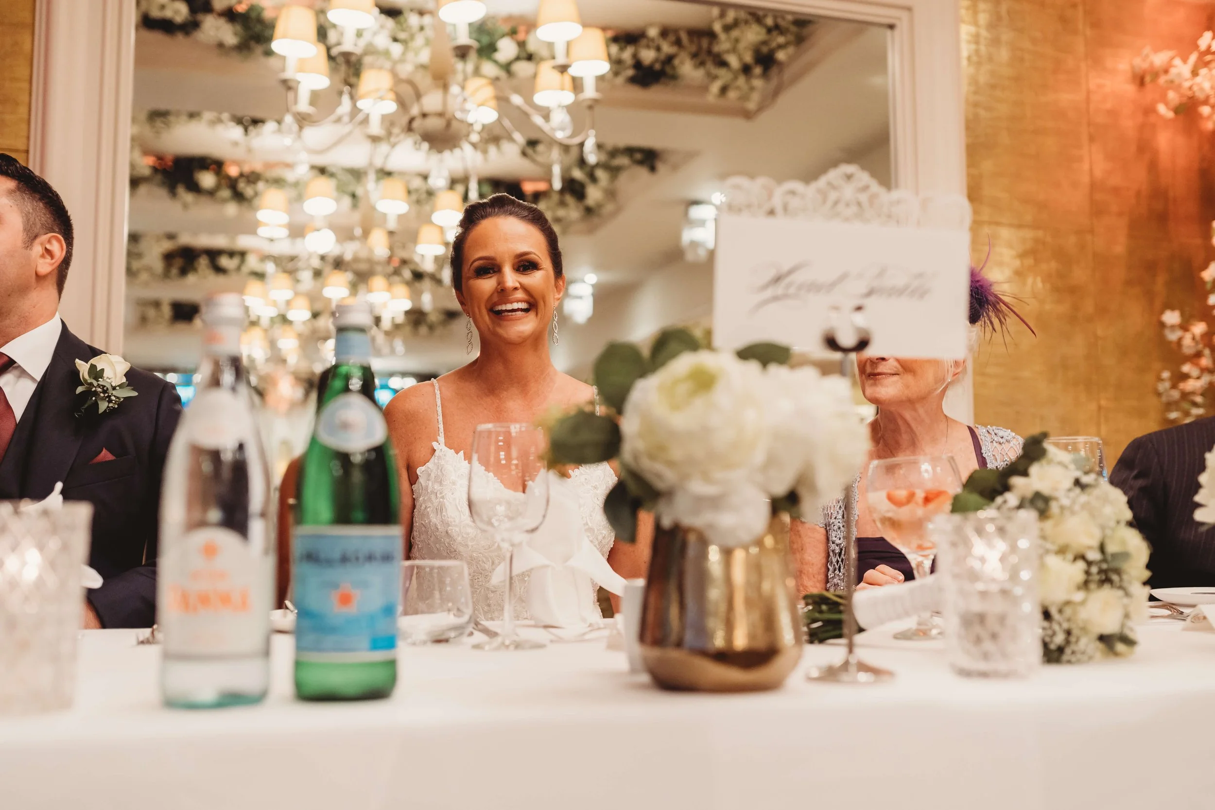 A smiling woman in a white dress sitting at a decorated table during a wedding reception. The table has floral centerpieces, a glass of water, and bottles of San Pellegrino and sparkling water. There are other guests partly visible around her, and th