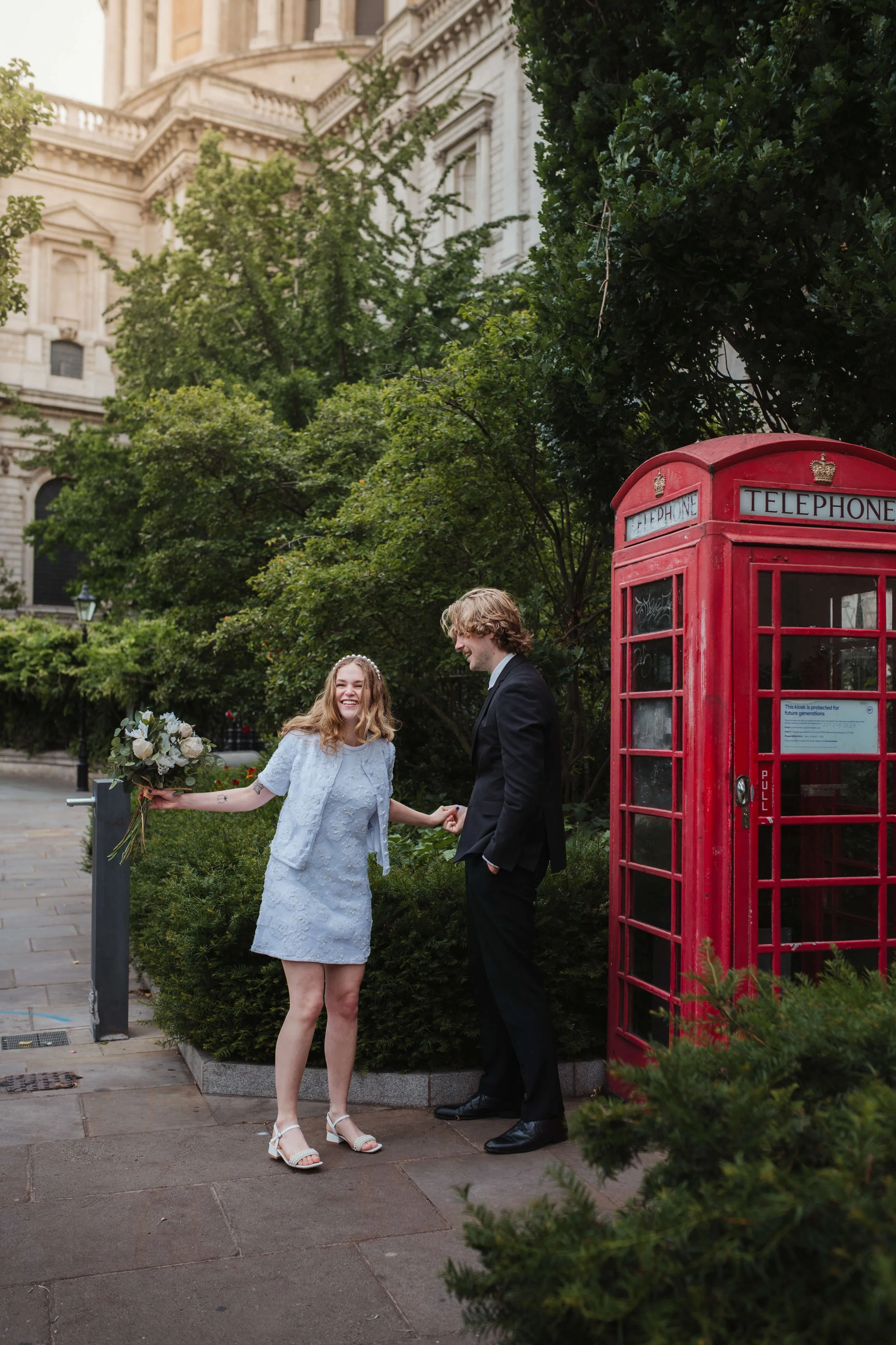 A young woman in a light blue dress holding a bouquet of flowers, smiling and holding hands with a young man in a black suit, standing next to a red telephone booth in an urban park setting.