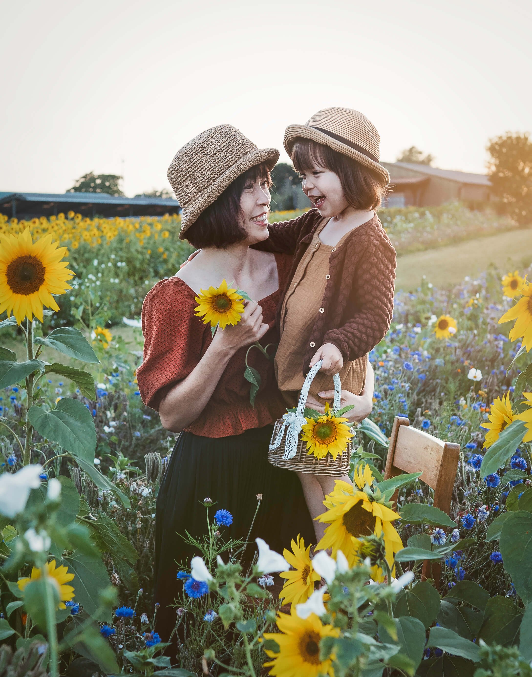 A woman and a young girl outdoors in a sunflower field, smiling and holding sunflowers, with the woman carrying a small basket decorated with sunflowers.