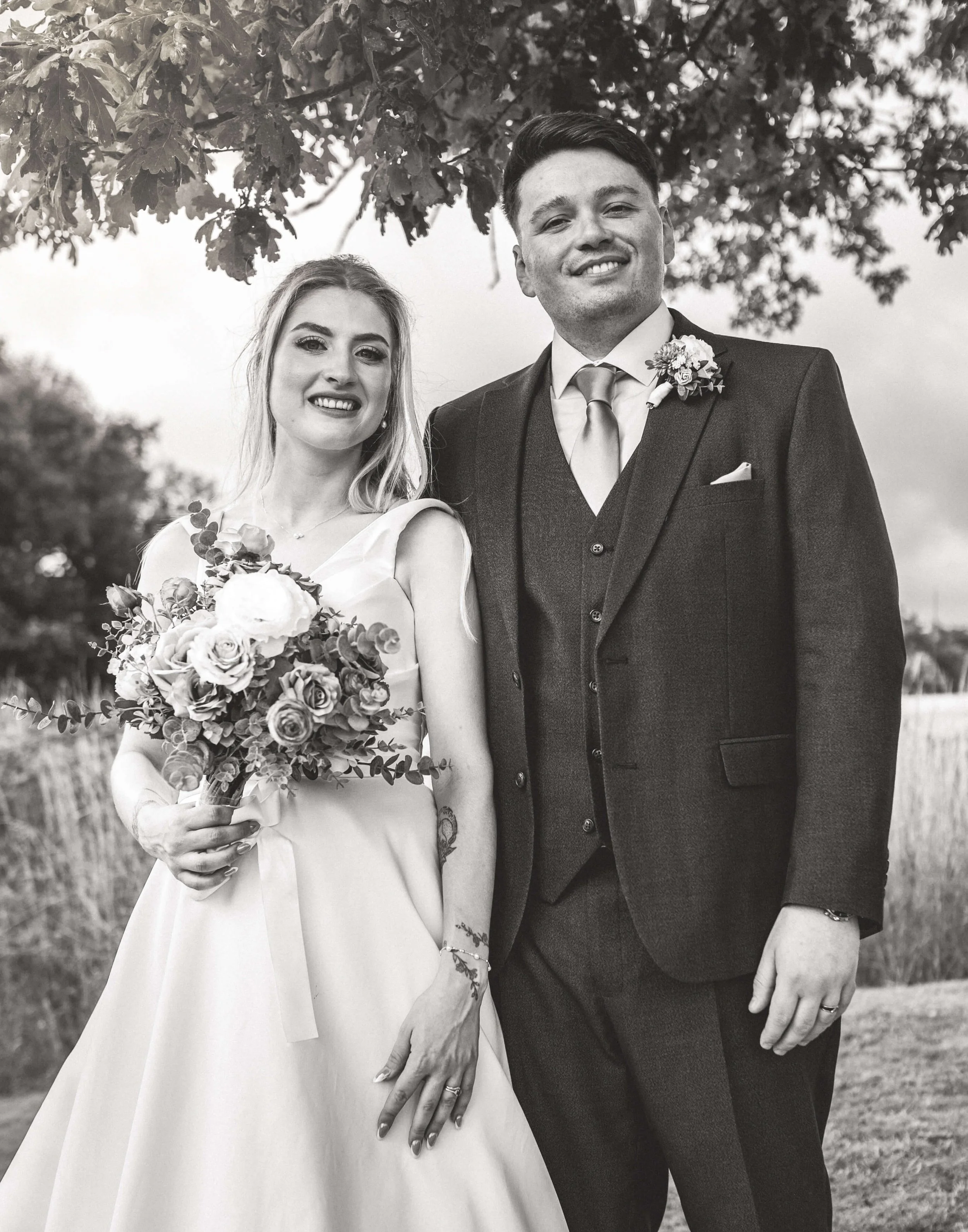 A black-and-white photo of a smiling bride and groom outdoors, standing under a tree on their wedding day. The bride is holding a bouquet of flowers and wearing a sleeveless wedding dress. The groom is dressed in a dark suit with a boutonniere on his