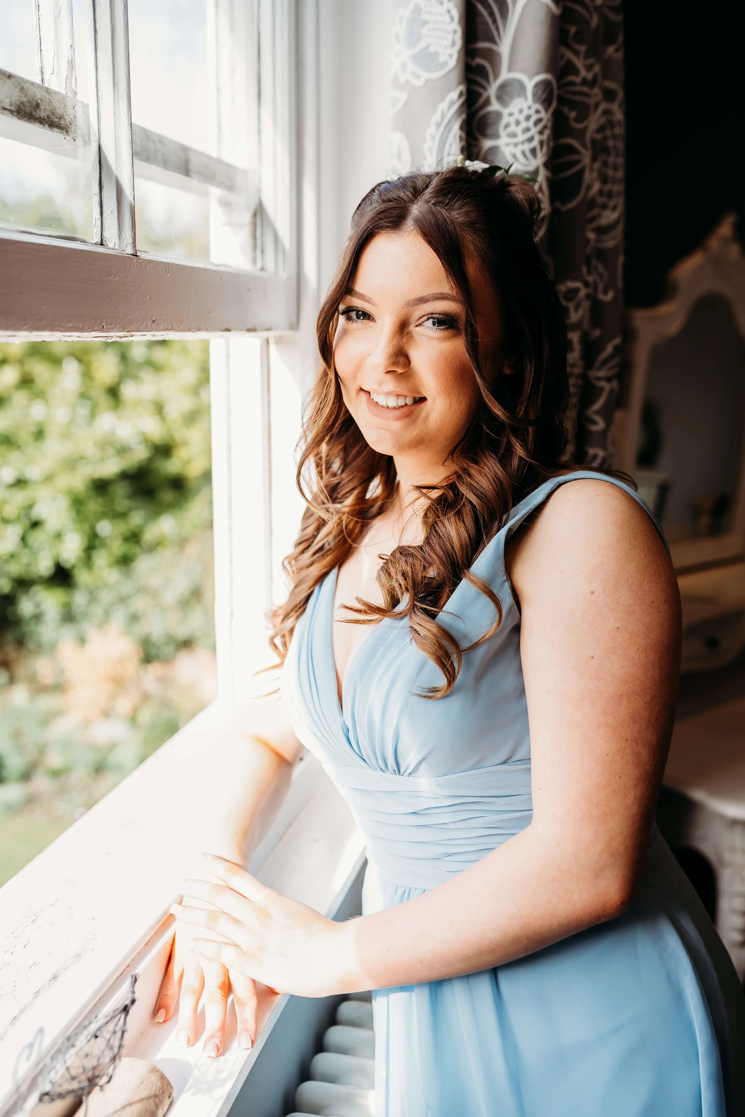A smiling young woman with long brown hair in loose curls, wearing a light blue sleeveless dress, standing near a window with sunlight streaming in. She is looking at the camera and resting her hand on a windowsill inside a room decorated with patter