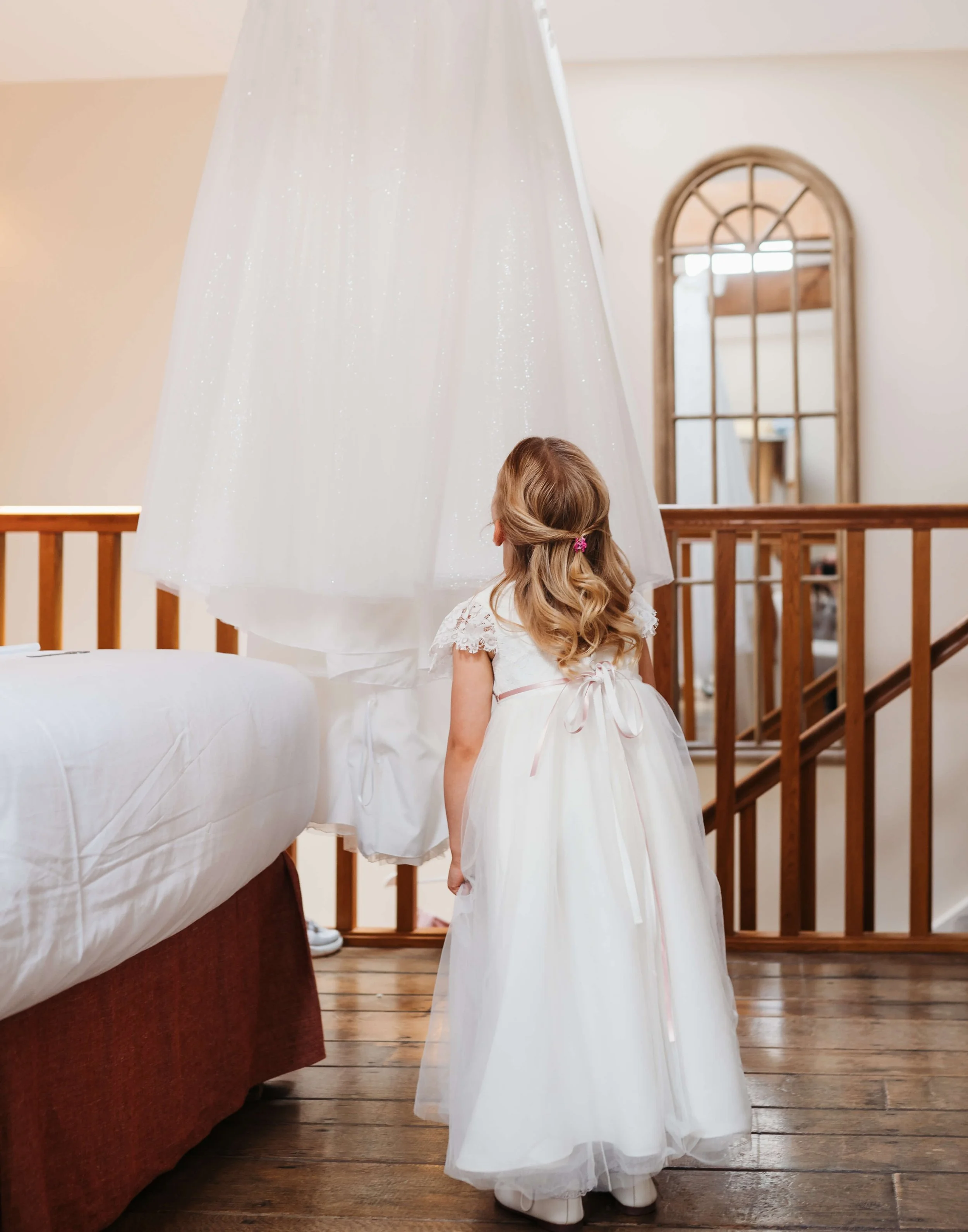 A young girl in a white dress with lace sleeves and a pink ribbon standing indoors, facing away, looking up at a hanging white canopy or curtain.