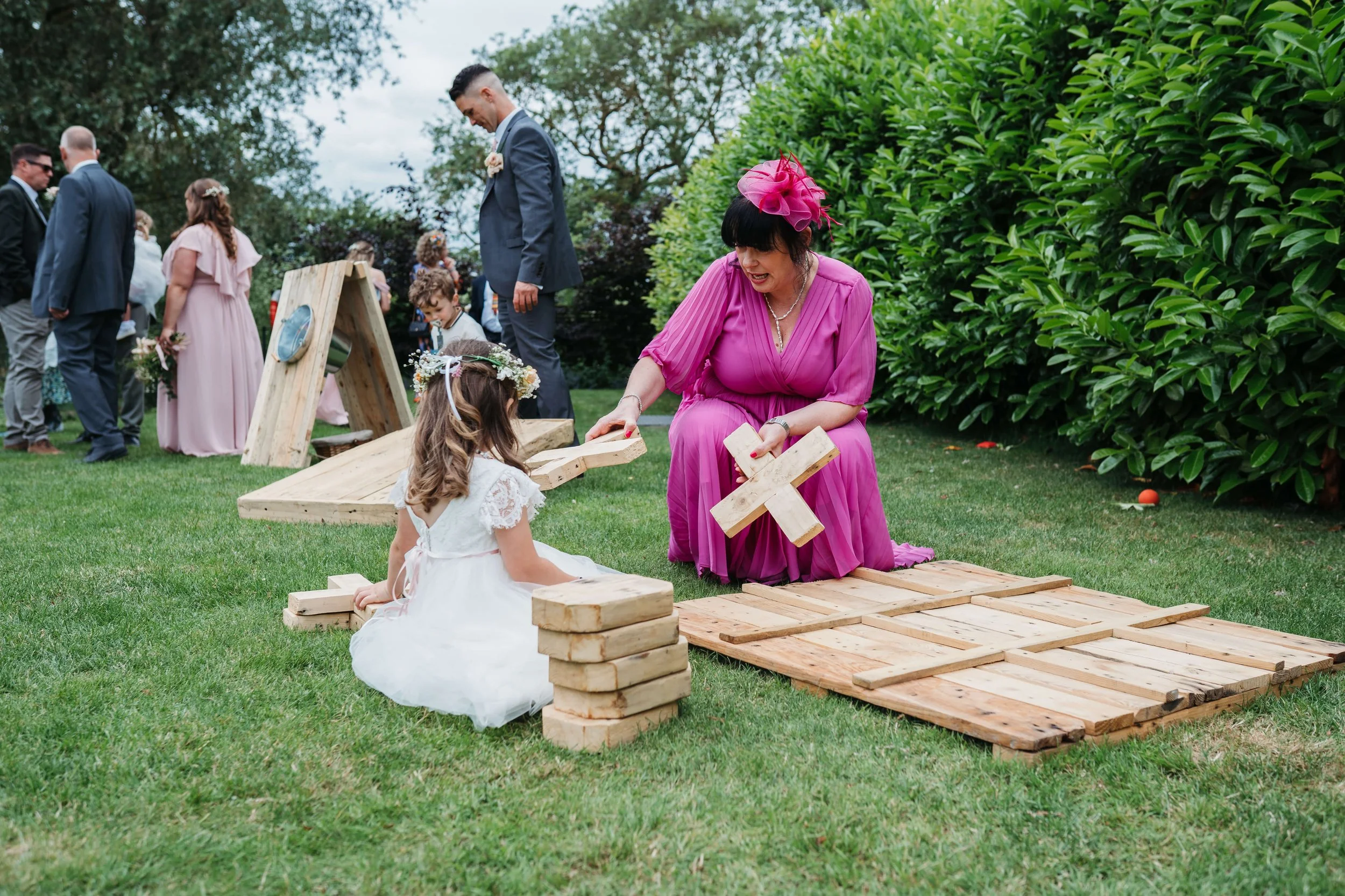 A woman in a pink dress and hat kneeling on the grass, helping two young girls build a wooden structure using large wooden blocks at an outdoor wedding celebration.