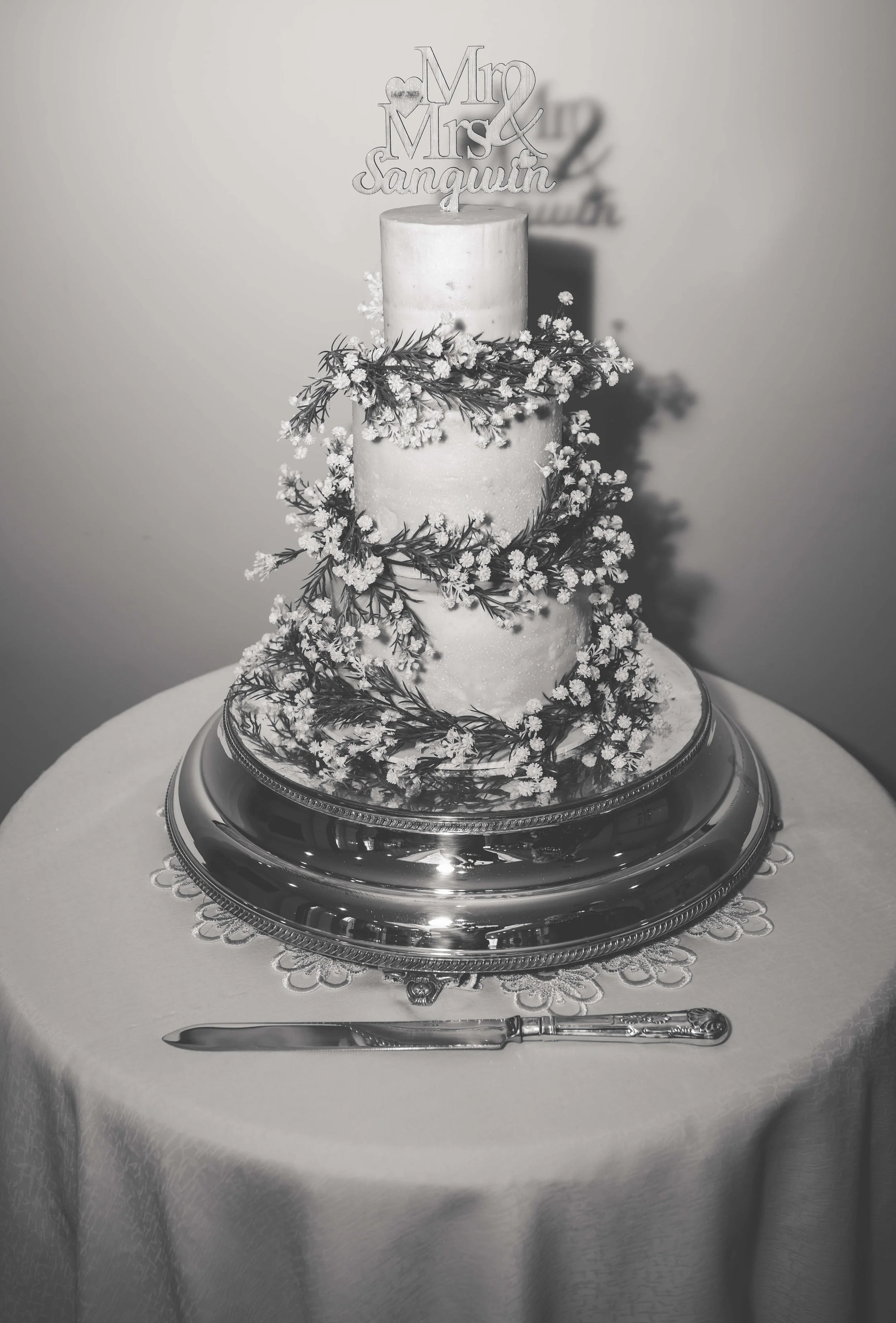 A three-tier wedding cake decorated with flowers and foliage, with a cake topper that reads 'Mr & Mrs Sanquilin,' on a table with a silver cake stand and a cake knife.