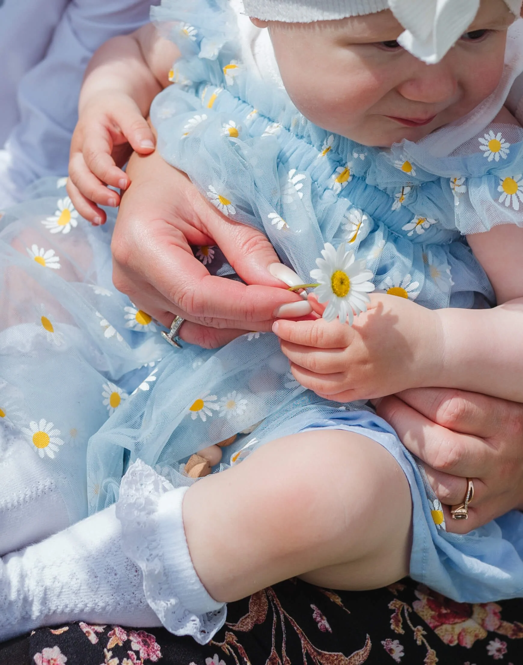 A baby girl dressed in a daisy-patterned blue dress receiving a daisy from an adult's hand.