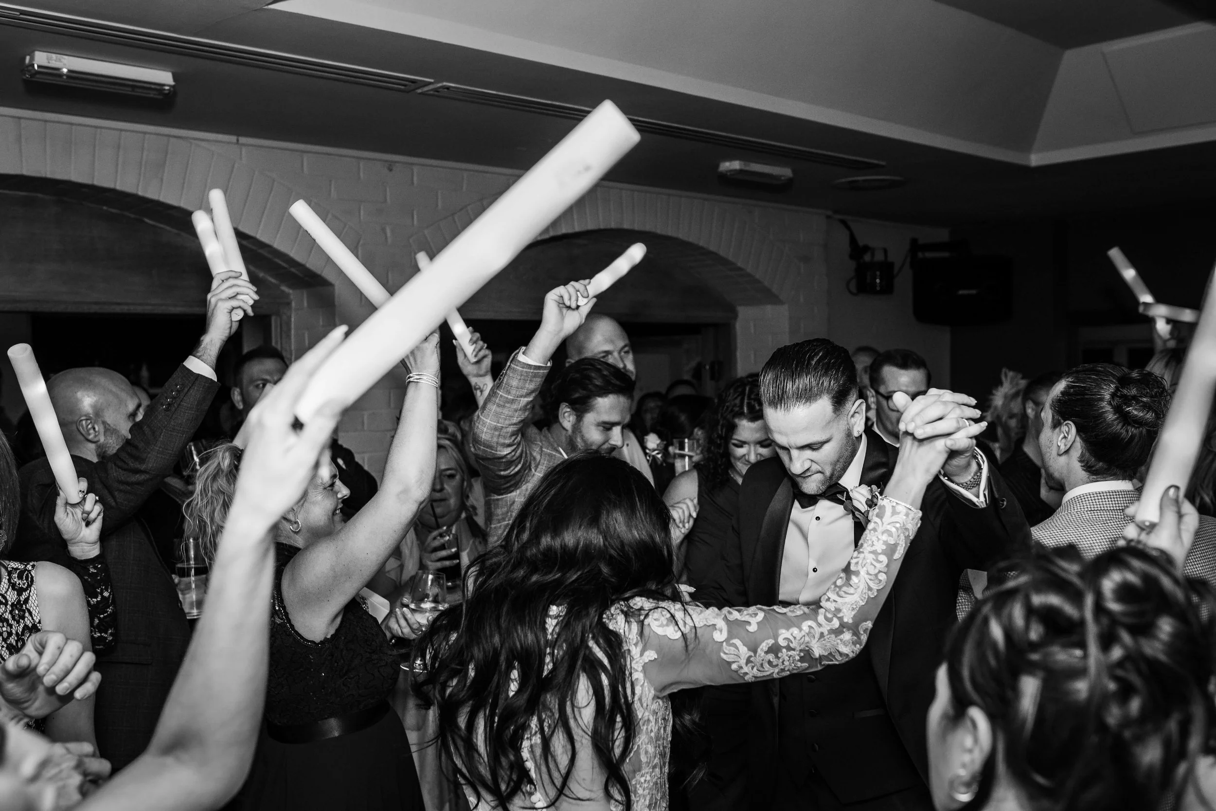 Black and white photo of a wedding reception with guests dancing and holding glow sticks. The bride and groom are dancing together in the center, surrounded by guests celebrating.