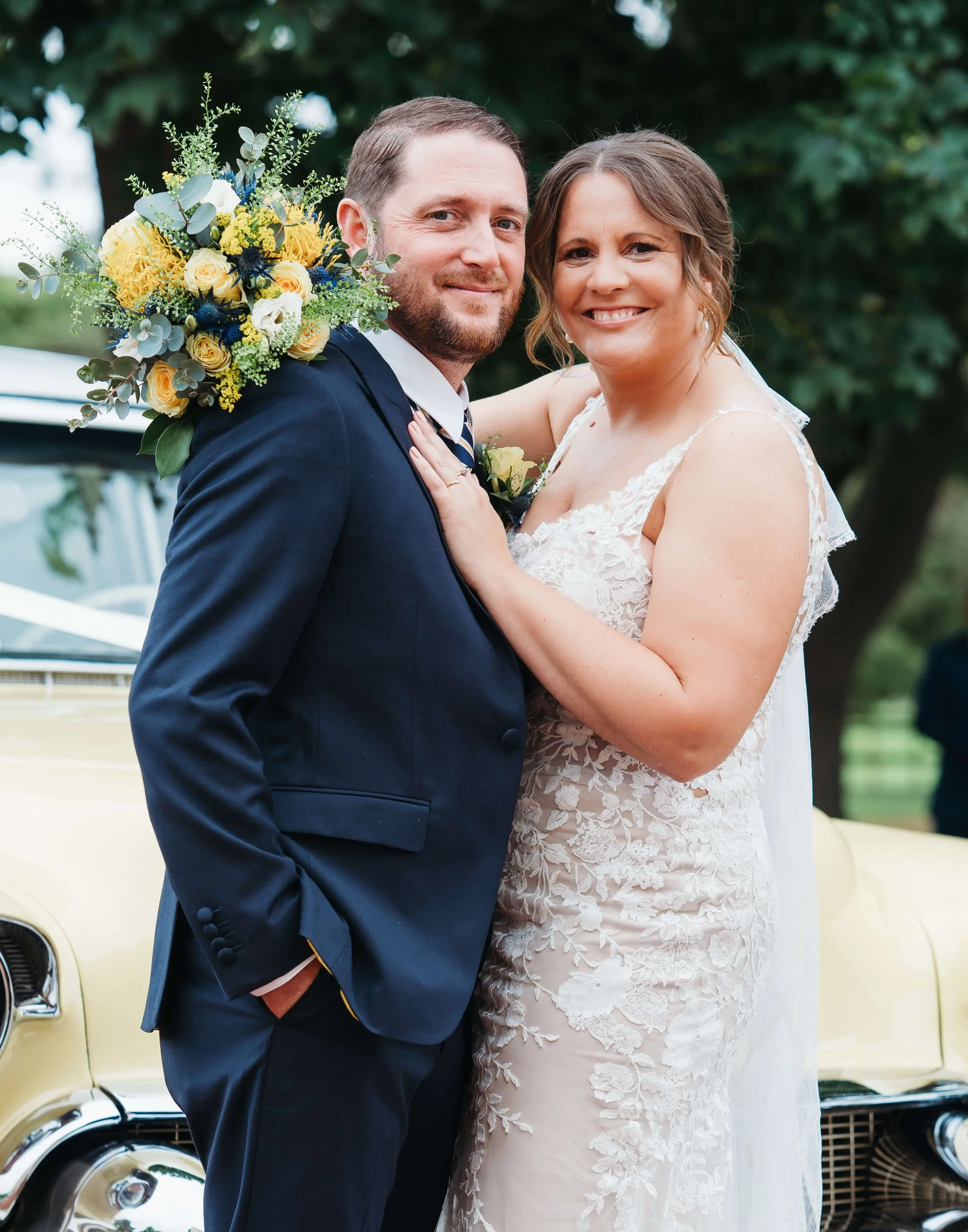 A newlywed couple smiling and posing outdoors, with the bride wearing a white lace gown and the groom in a navy suit, holding a bouquet of yellow and blue flowers.
