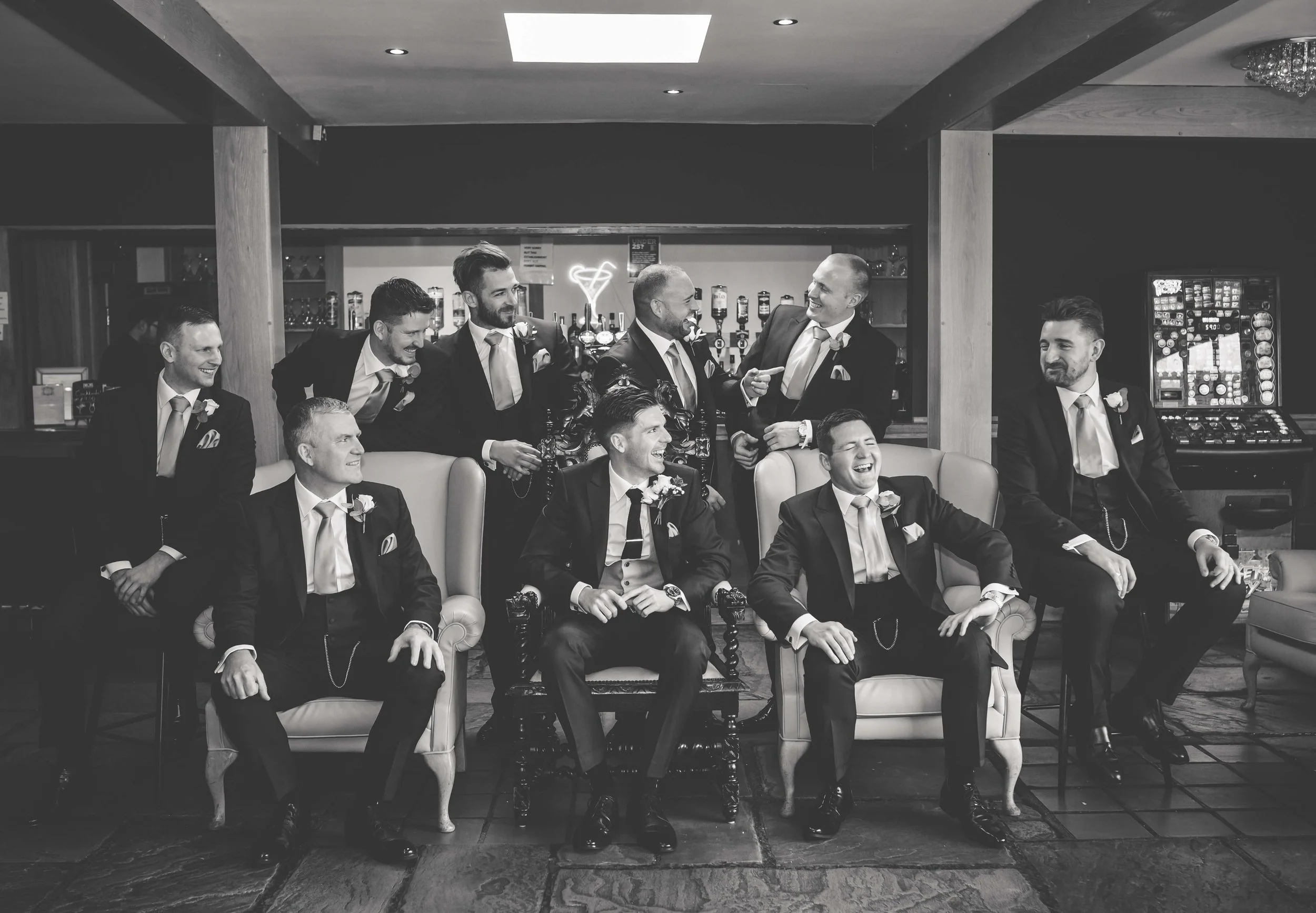 Black and white photo of ten men in suits, some seated and some standing, smiling and laughing in a bar or lounge setting, with a neon heart sign in the background.
