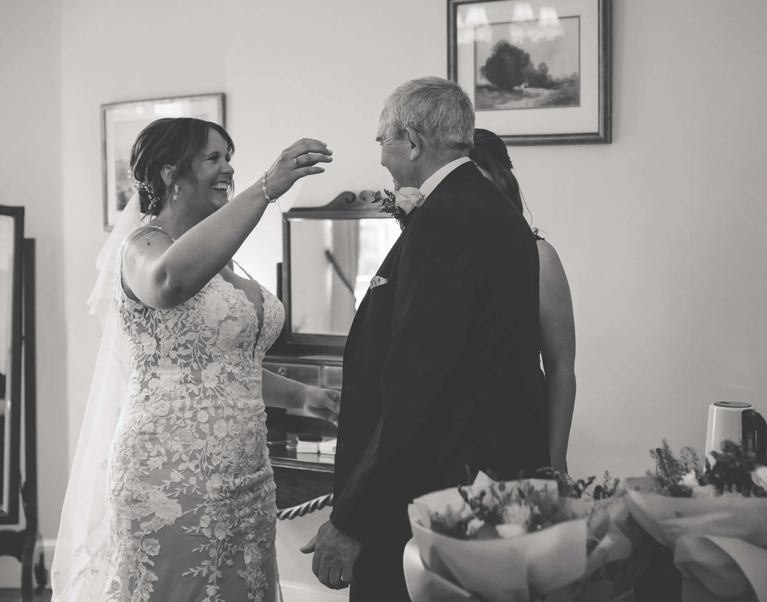 A bride giving a bouquet to an older man, possibly her father, during a wedding celebration. The scene is indoors with framed artwork on the wall and floral arrangements on the table.