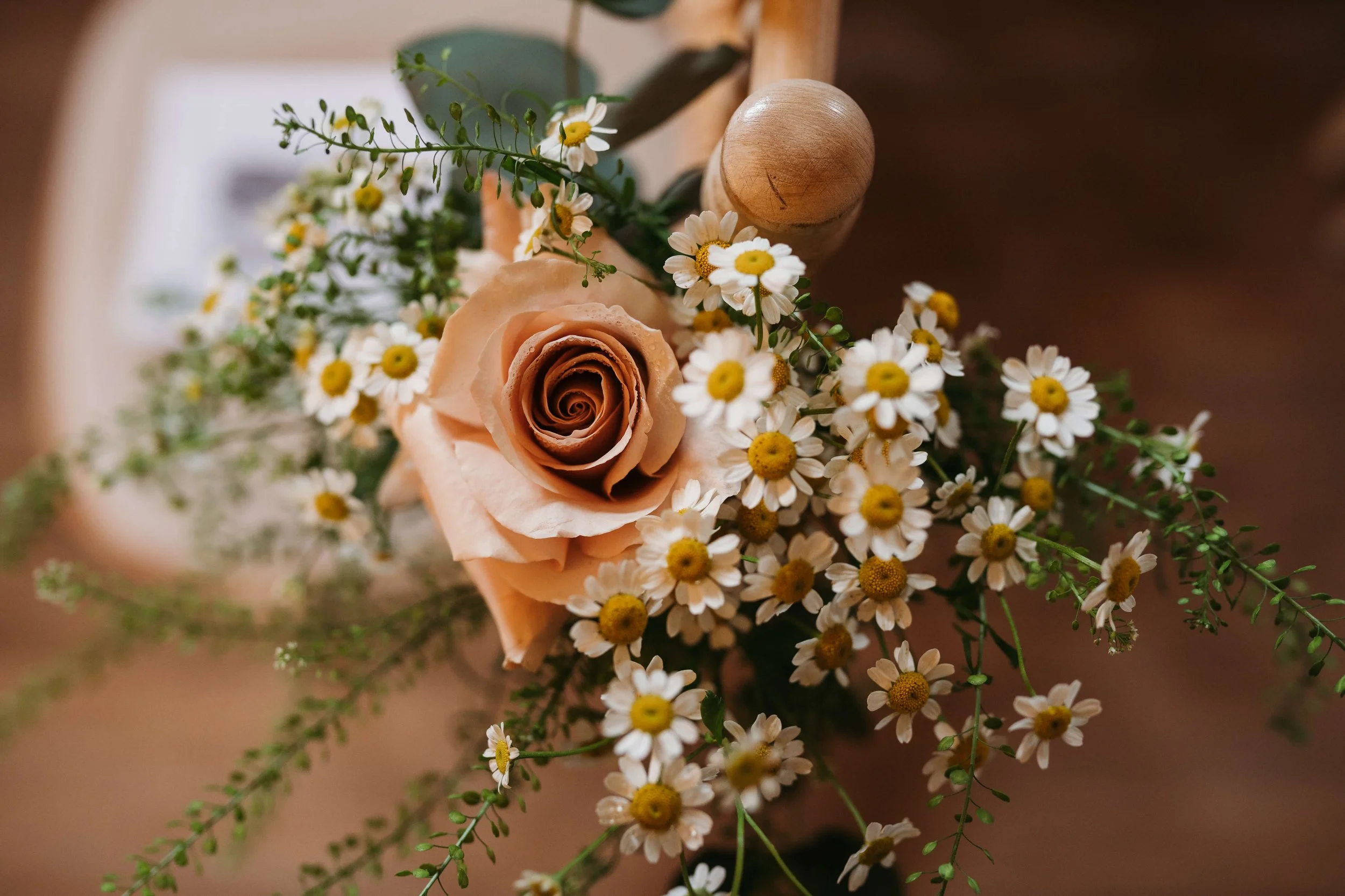 A close-up of a small bouquet of flowers featuring a peach-colored rose surrounded by small white daisies with yellow centers, green leaves, and filler greenery.