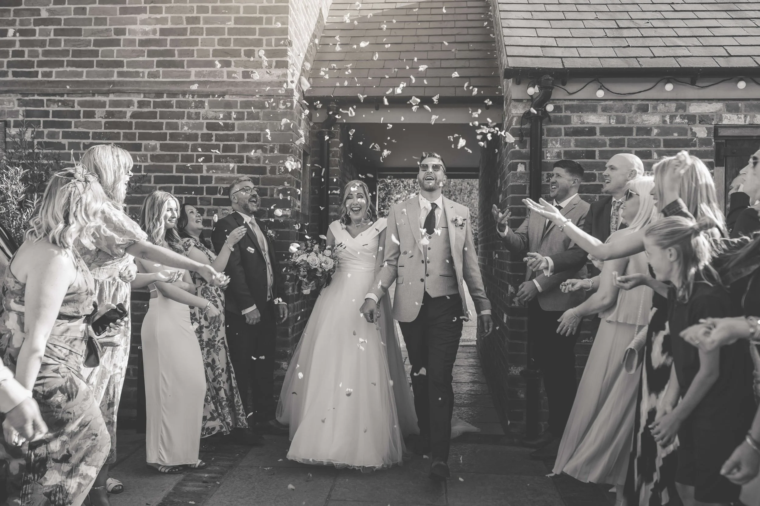 A bride and groom walking out of a brick building while friends and family celebrate and throw confetti.