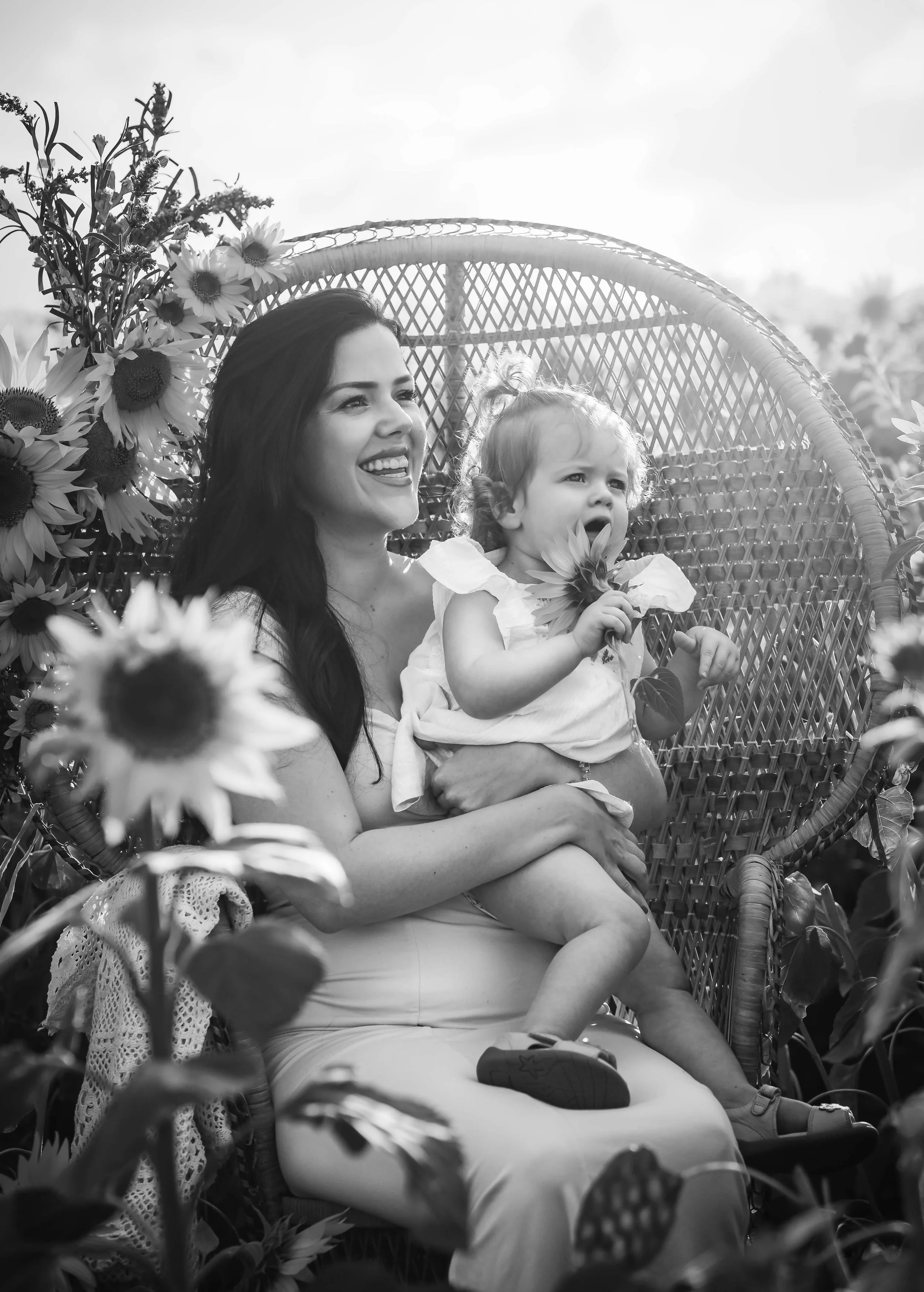 A woman with long dark hair and a young girl with curly hair sitting on a decorative chair surrounded by sunflower plants. The woman is smiling and the girl is holding flowers, with an outdoor setting in the background.