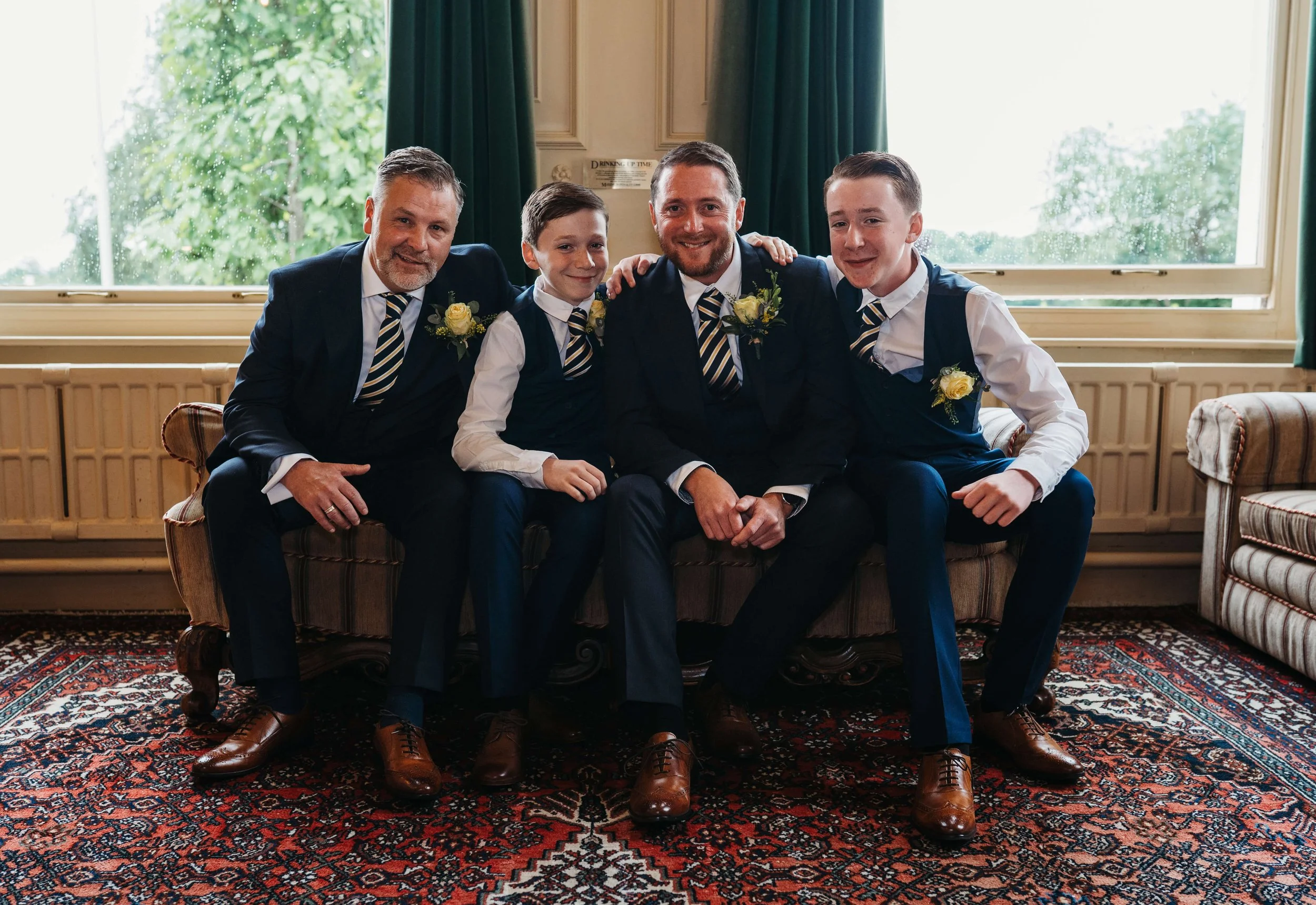 A group photograph of four men and boys in formal attire sitting on a vintage sofa in a room with large windows and a patterned rug. The men are wearing dark suits, white shirts, striped ties, and boutonnieres, while the boys are dressed in navy vest