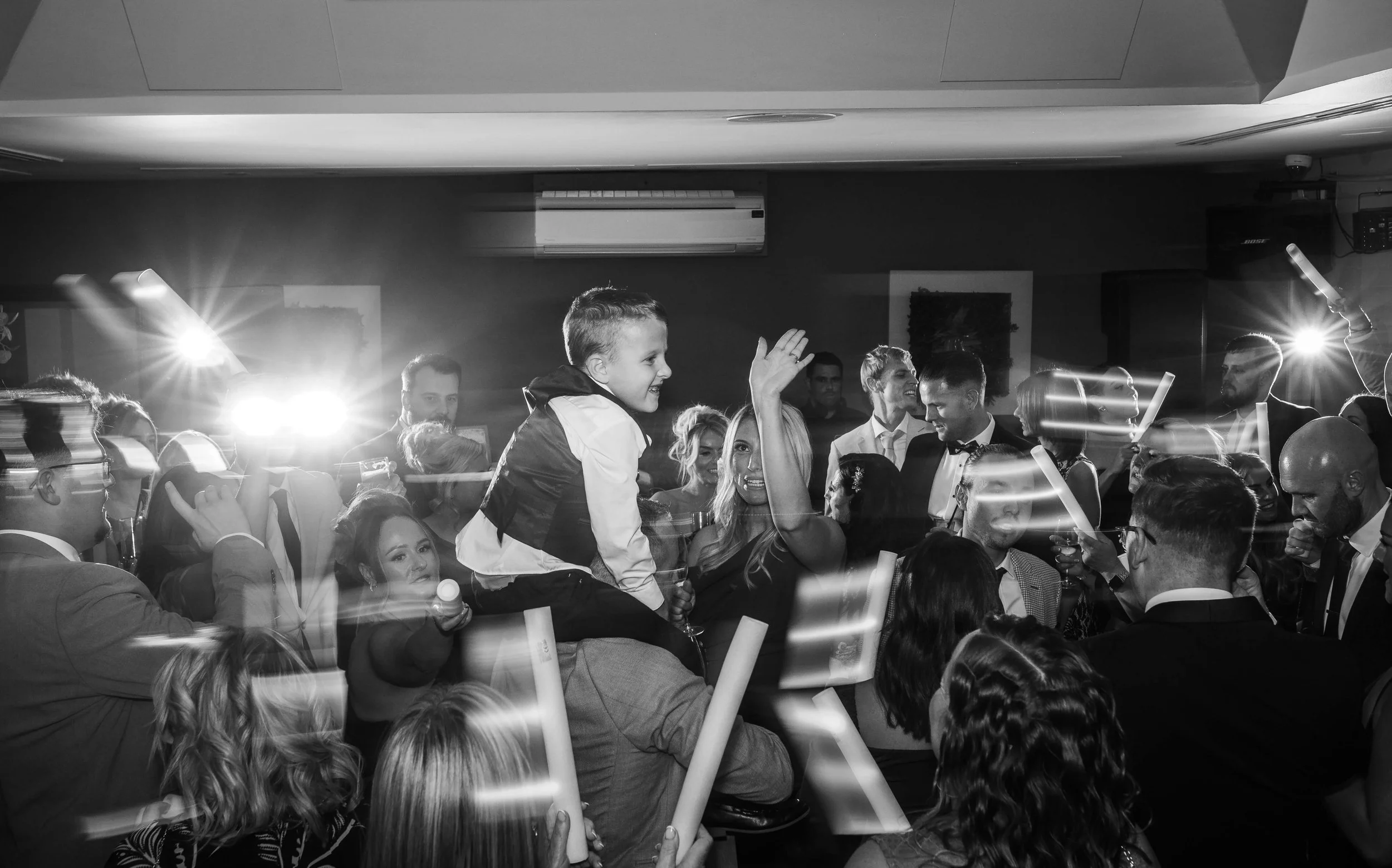 A black and white photo of a lively celebration with many people dancing and cheering, including a boy sitting on a woman's shoulders, raising his hand, with bright lights and an energetic atmosphere.
