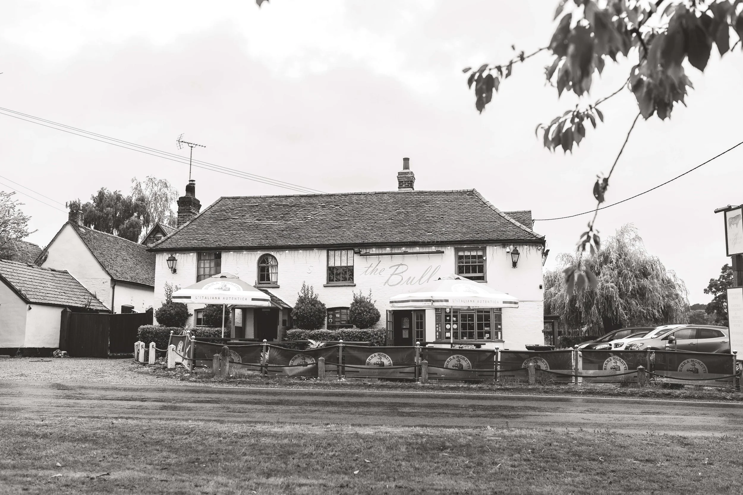 Black and white photo of a two-story pub called 'the Bull' with outdoor seating and umbrellas, cars parked nearby, and trees in the background.