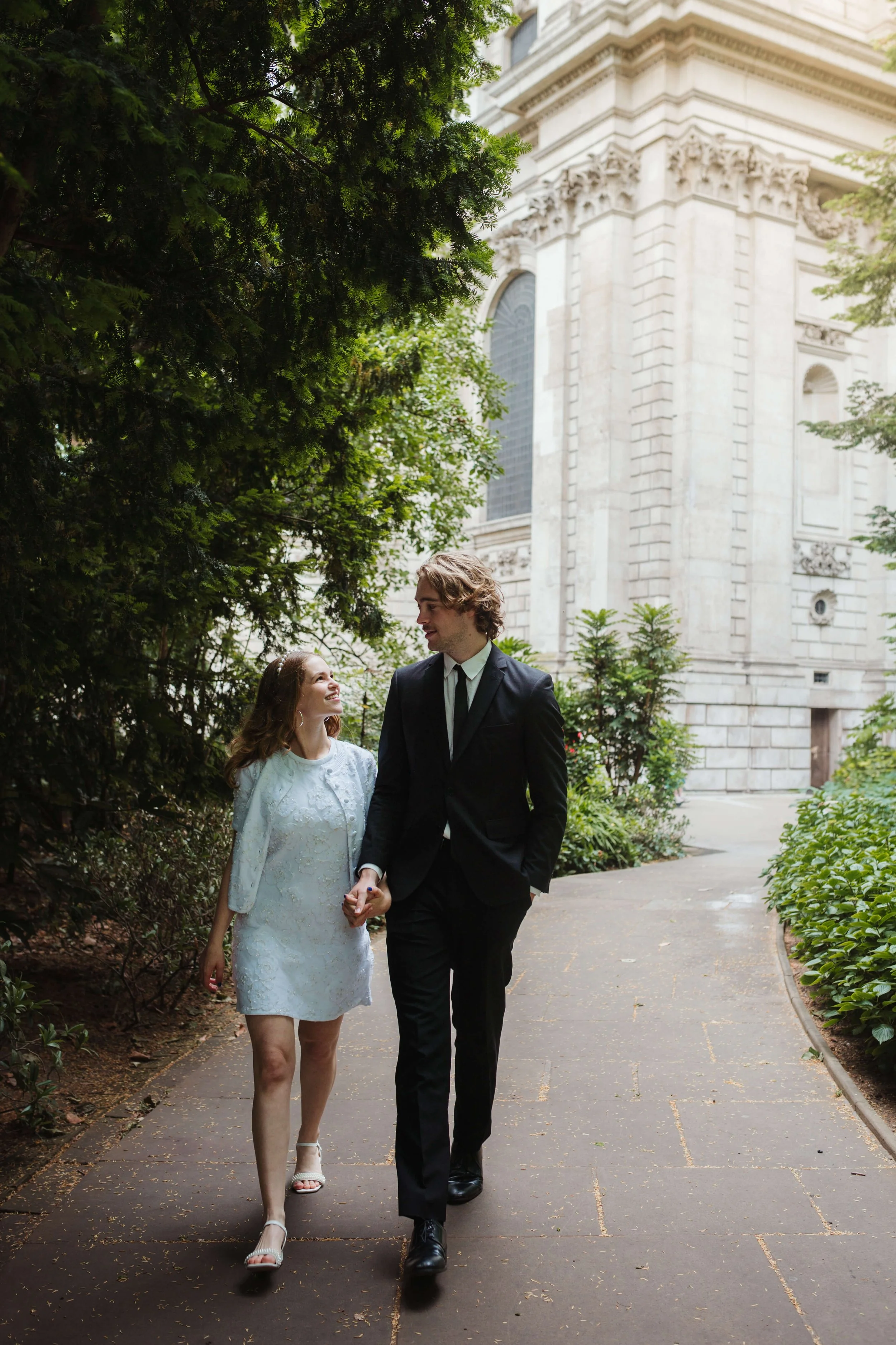 A young couple walking hand in hand on a path surrounded by greenery, with a historic stone building in the background.