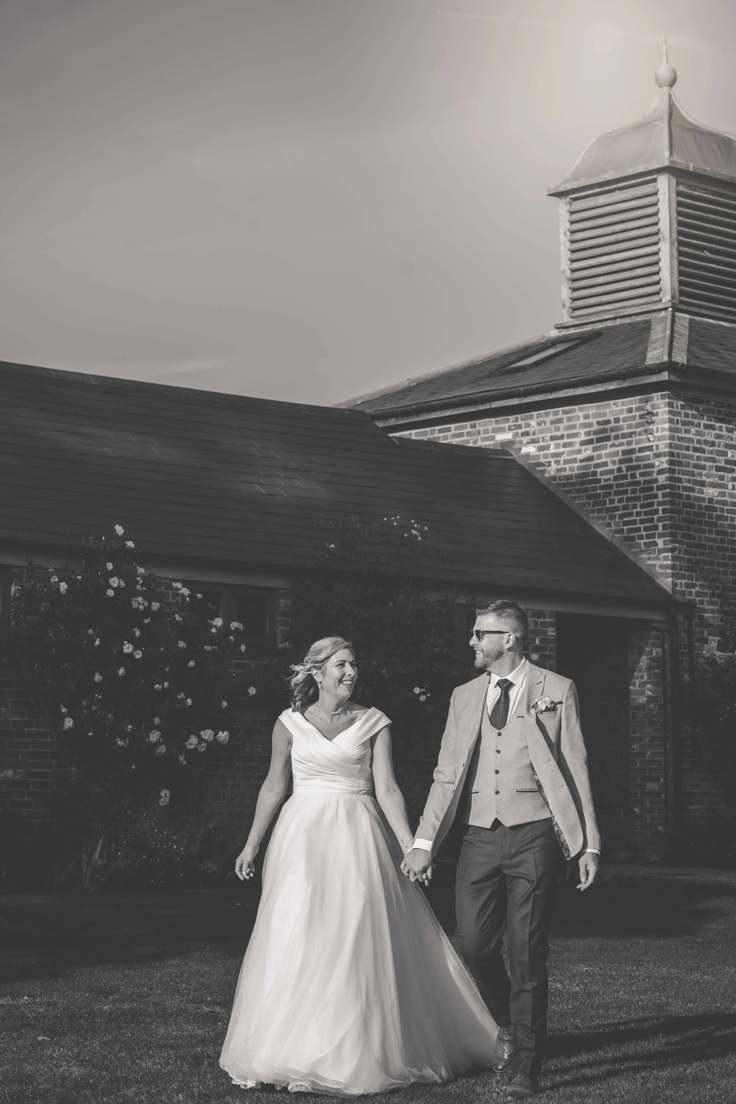 A black and white photo of a bride and groom walking hand in hand outside a brick building with a steeple, smiling and looking at each other.