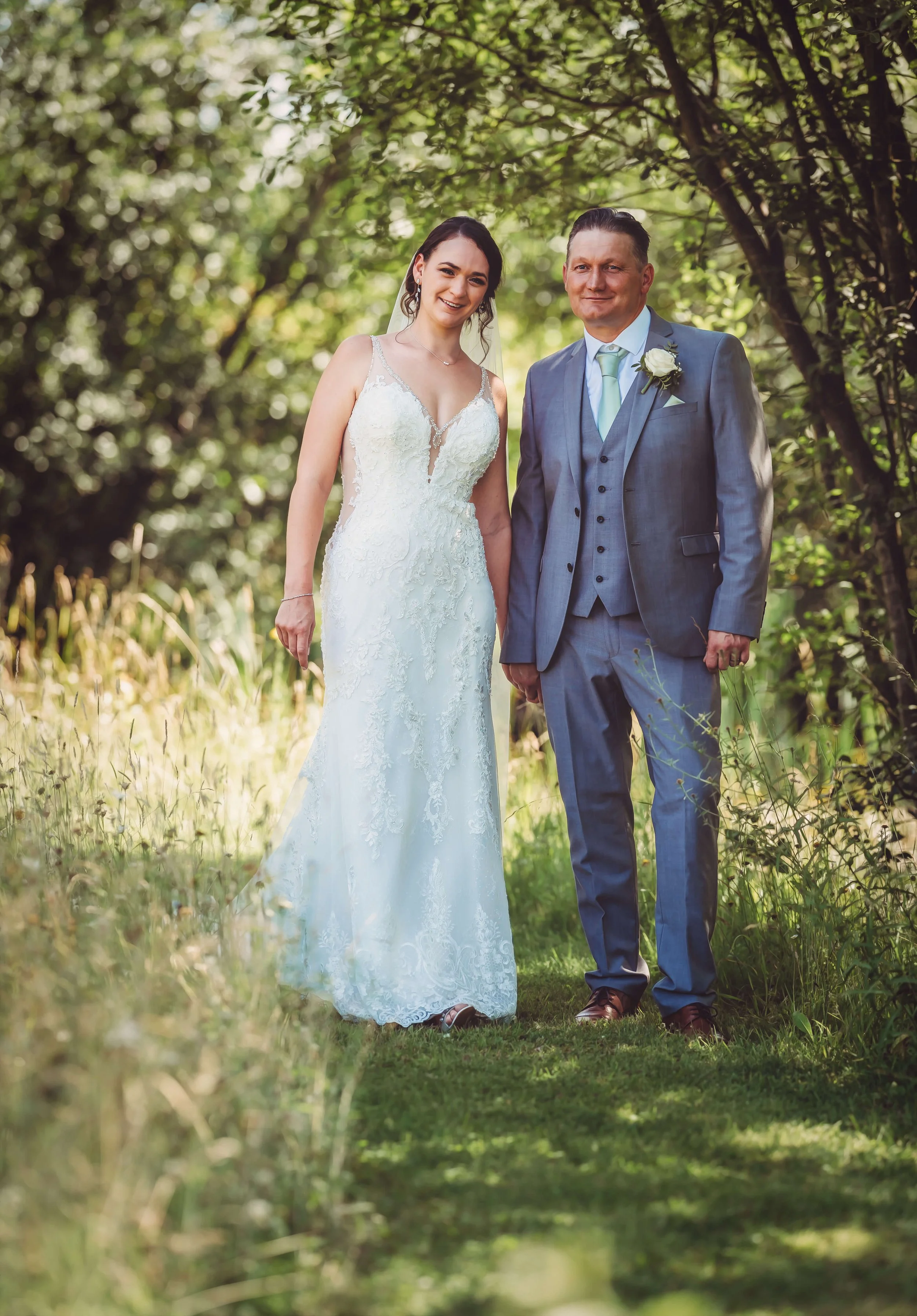 A bride and a groom standing together outdoors in a wooded area on their wedding day, smiling at the camera.